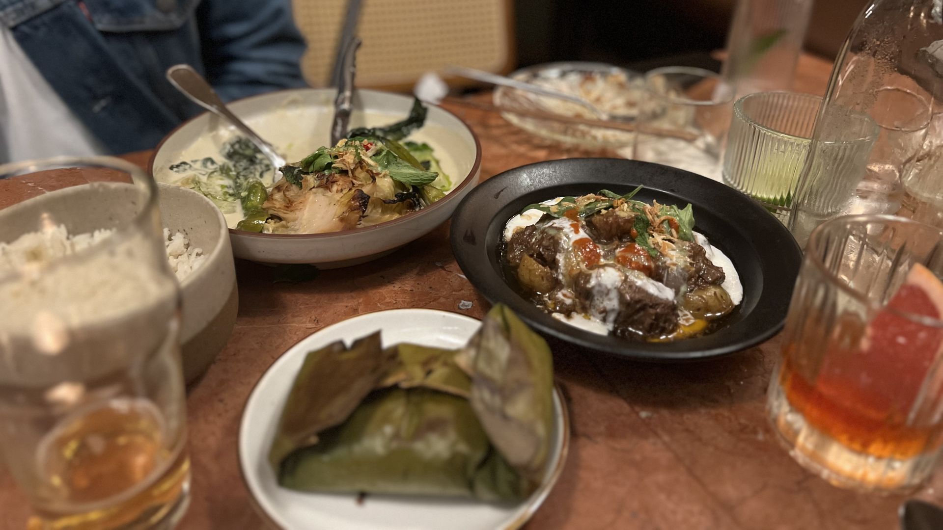Table with various dishes including a creamy vegetable dish, a black bowl with meat and sauce, wrapped food on a white plate, glasses with drinks, and a person in a denim jacket.