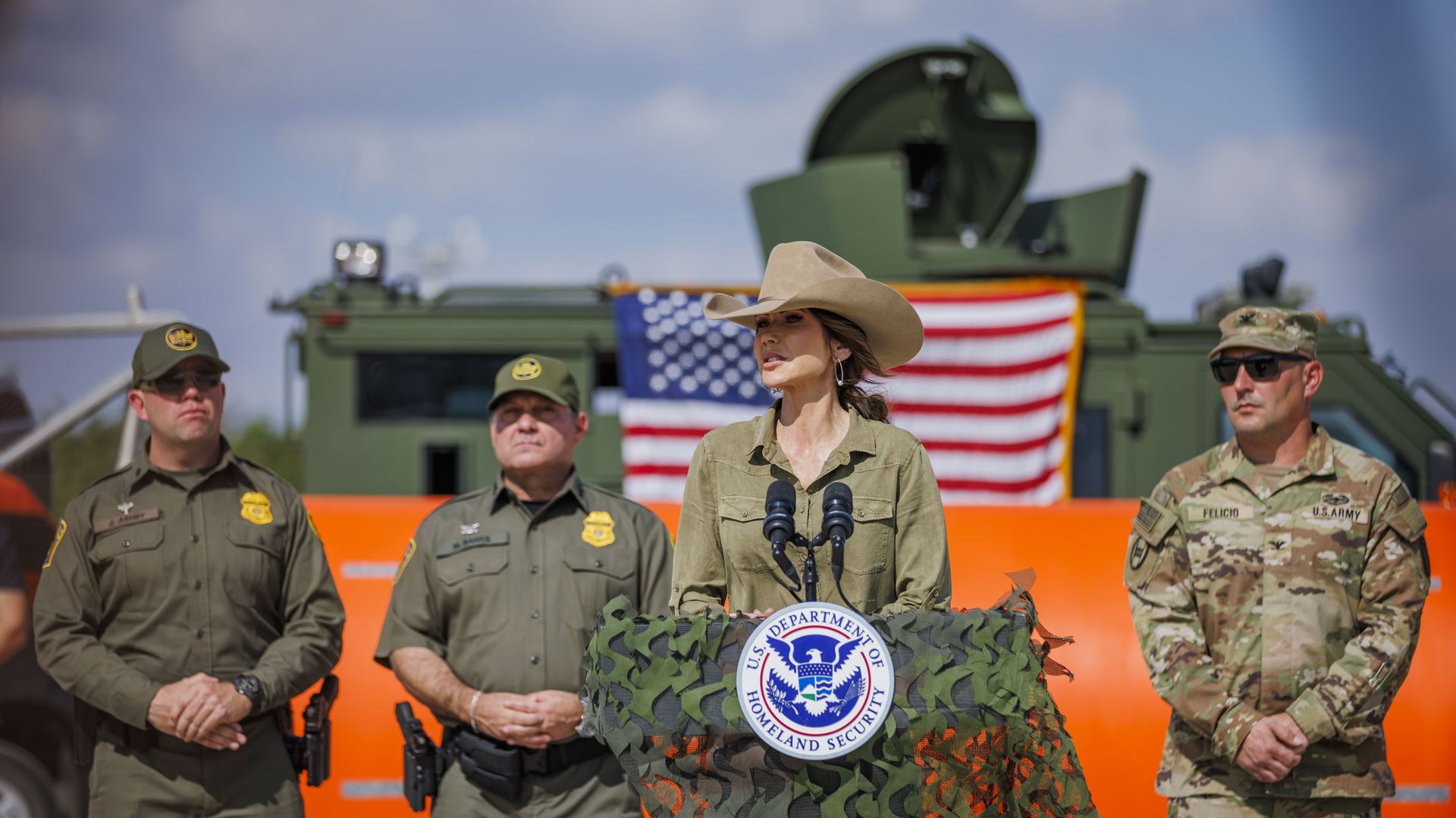 Kristi Noem — wearing a khaki cowboy hat and button-down shirt — speaks into microphones attached to a podium covered in green fabric while uniformed officials stand behind her.