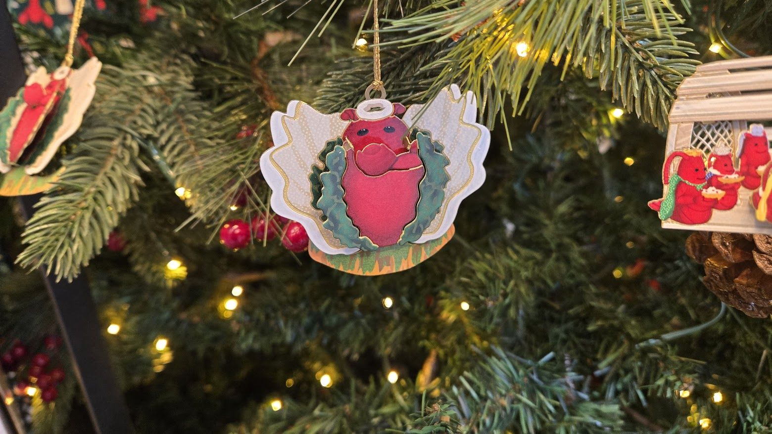 Christmas tree with lights and red berry decorations, featuring an angel ornament shaped like a red lobster with a halo, white wings, and green holly leaves.