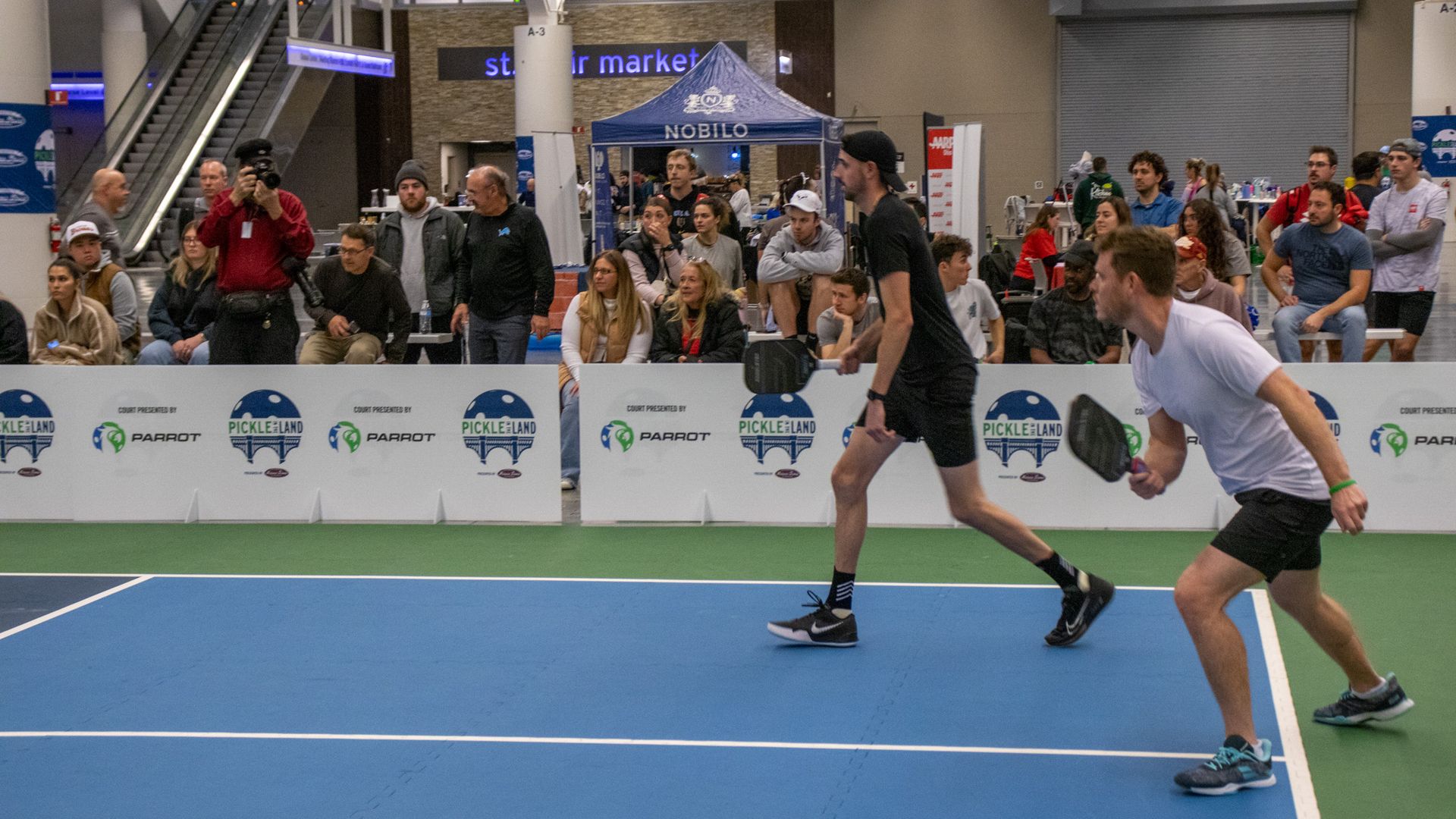Two men playing pickleball indoors on a blue and green court, one in black and one in white, with spectators and photographers behind a barrier branded with "Pickle Land" logos.