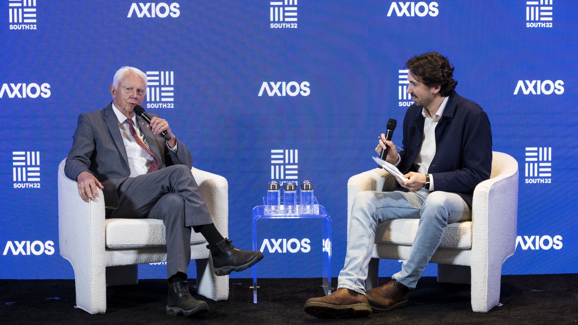 Two men sit on an event stage. The man on the left, a congressman, is in a suit. The man on the right, a reporter, is dressed more casually.