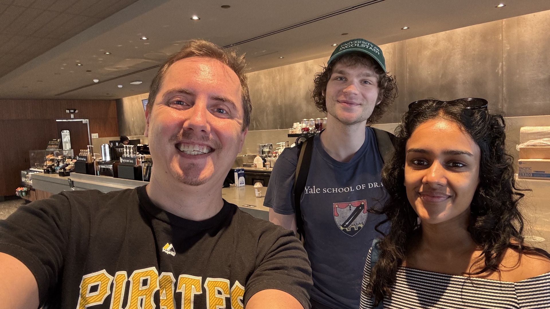 Three smiling people indoors in a coffee shop; man in black "PIRATES" shirt, man in blue "Yale School of Drama" shirt and green cap, woman with curly hair wearing striped top and sunglasses on head.