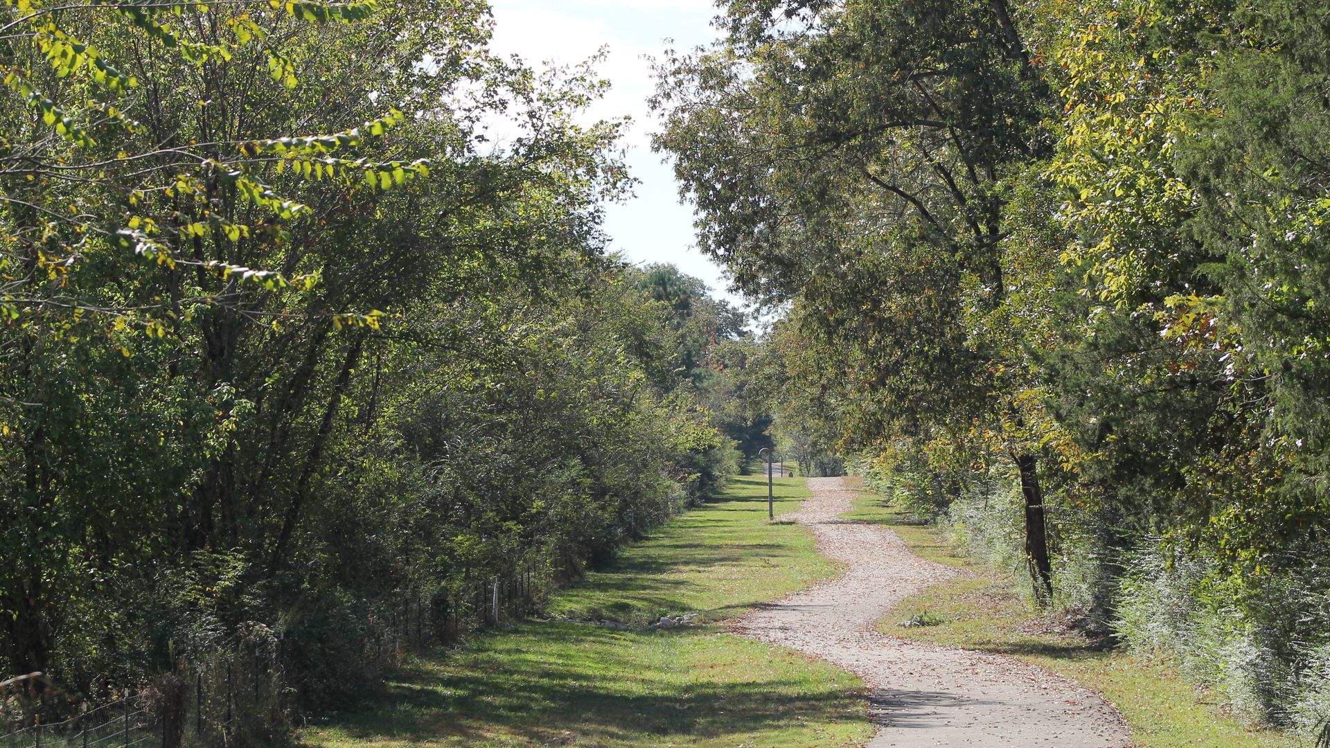 A winding gravel and pavement path surrounded by dense green trees and grass under a clear sky on a sunny day.