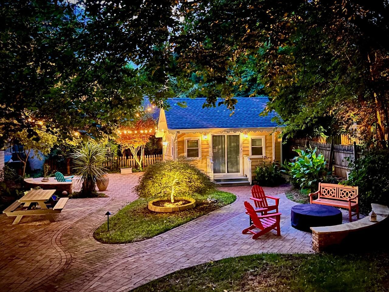 Cozy backyard at dusk with lit patio lights, a small yellow house, red chairs around a covered fire pit, a wooden bench, picnic table, and lush green trees.