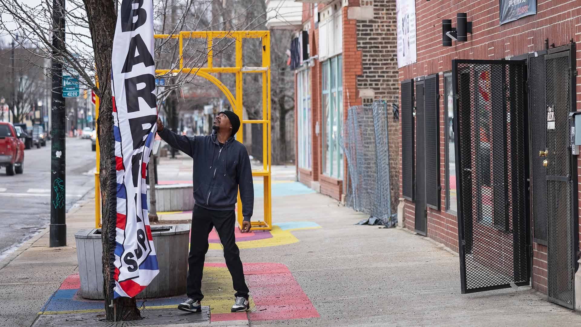 Man in a black hoodie and beanie adjusting a red, white, and blue barber shop flag attached to a tree on a city sidewalk with colorful painted squares and brick buildings.