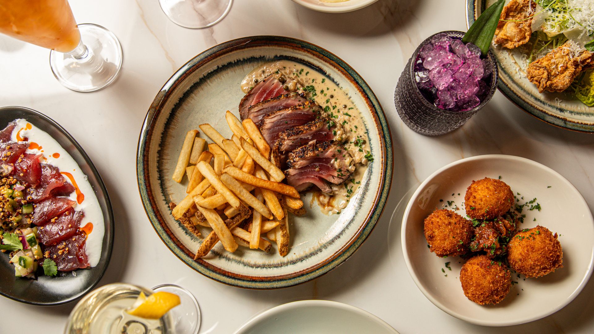 A table set with multiple dishes, including fritters, drinks and seared tuna.