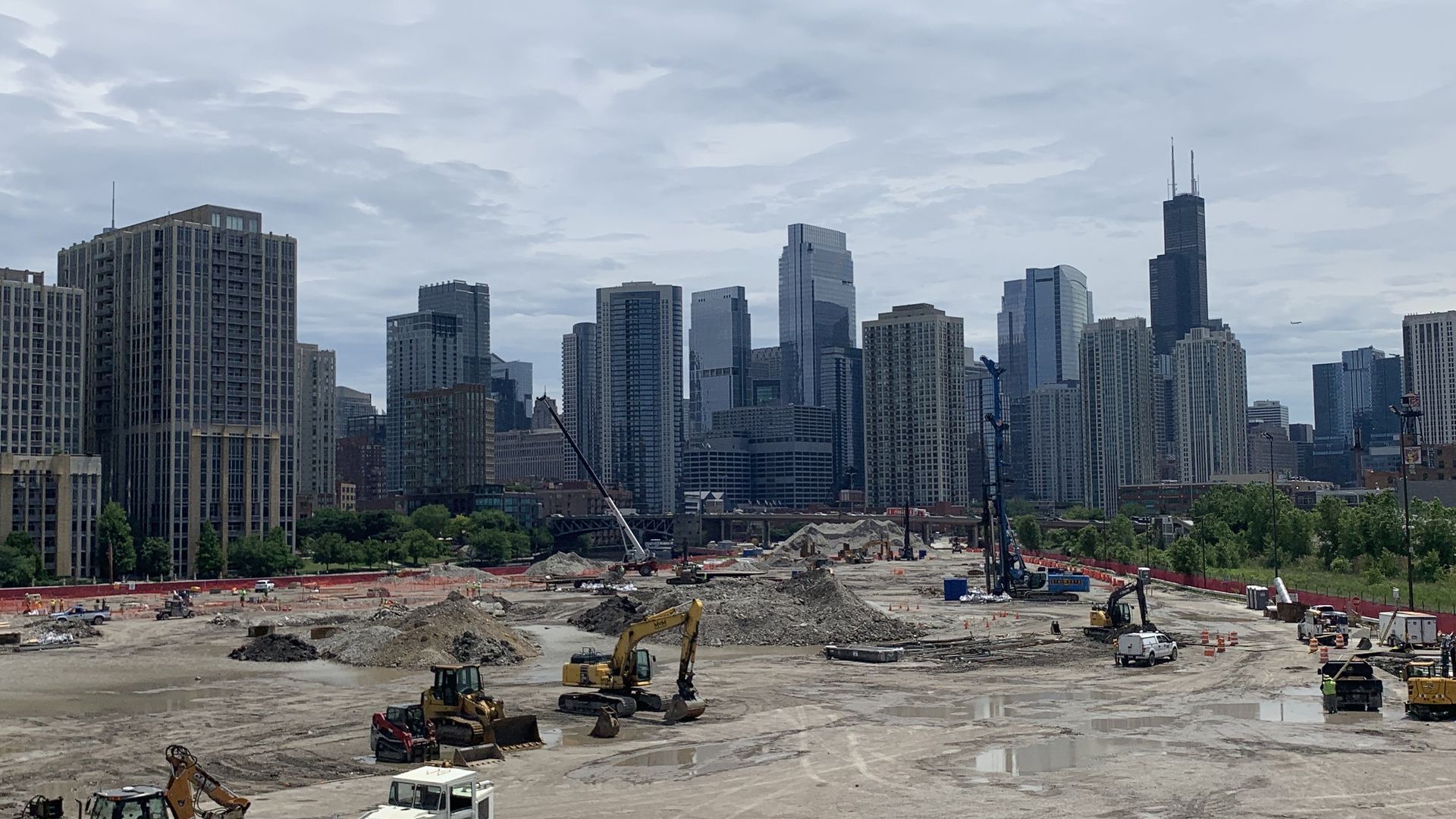 Construction site with heavy machinery and workers in front of a city skyline with tall buildings under a cloudy sky.