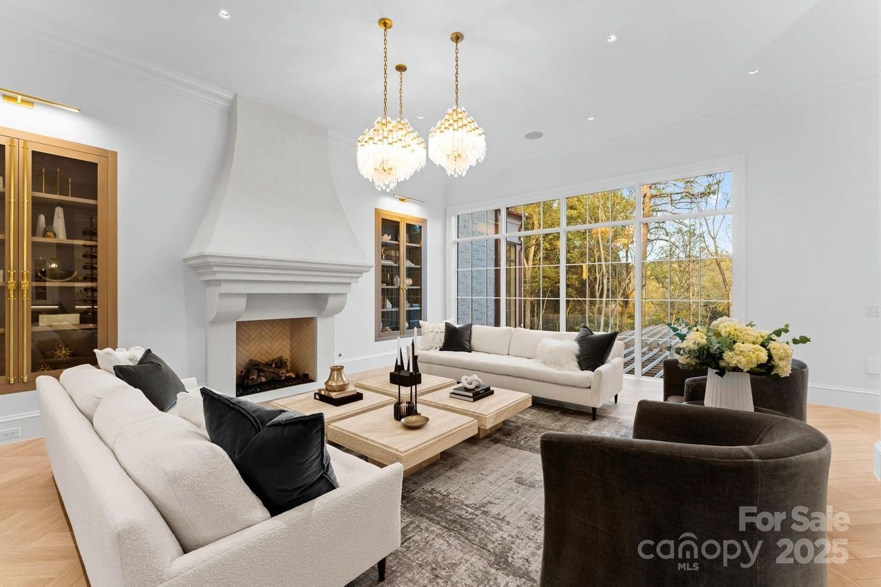 Bright modern living room with white sofas, dark armchairs, wooden coffee tables, large windows showing trees outside, white fireplace, gold light fixtures, and built-in cabinets.