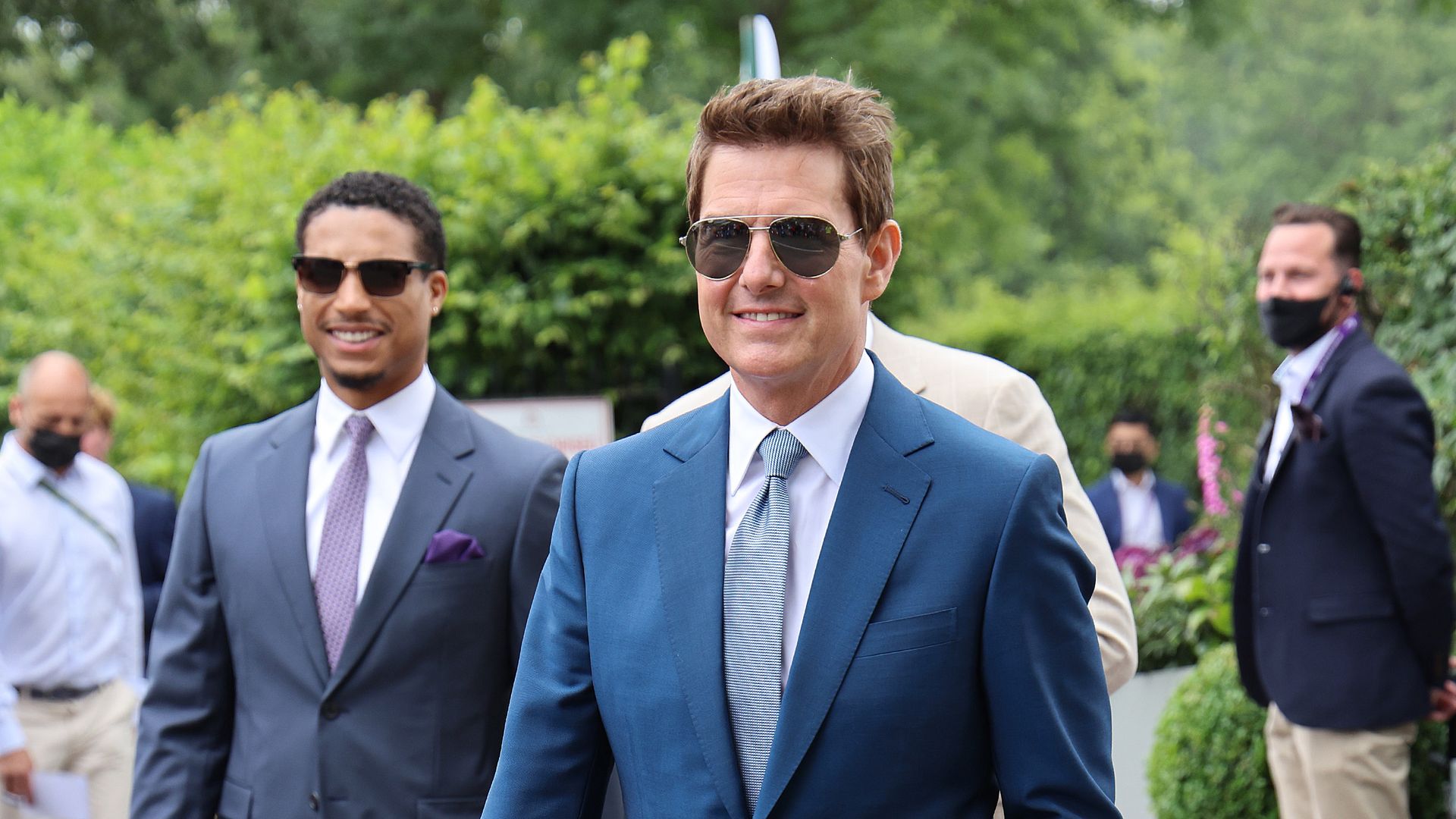 Photo of Tom Cruise in a navy blue suit and aviator sunglasses walking outside 