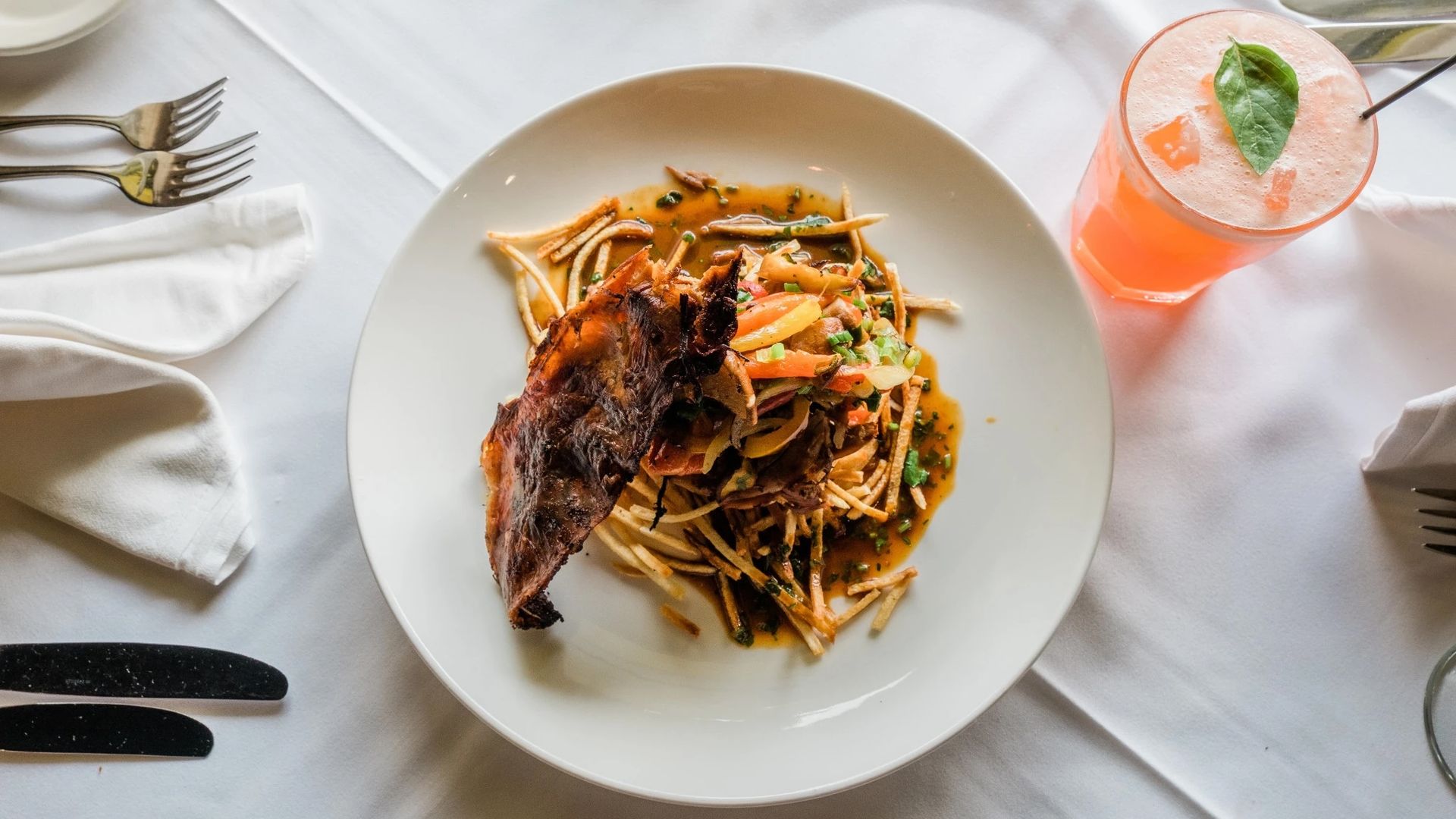 A plate of slow roasted duck sits on a white plate on a white tablecloth. Dining utensils sit to the left, and an orange-colored cocktail sits to the right.