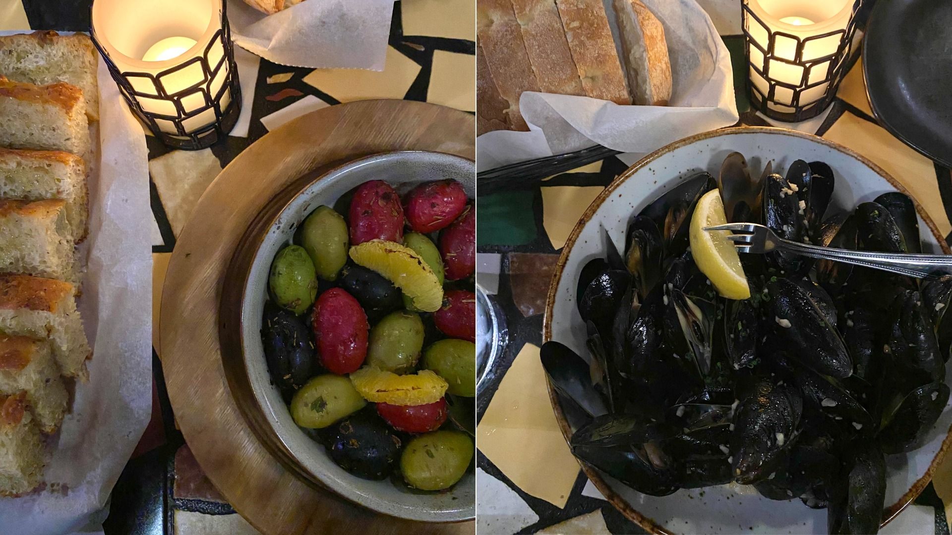 Dinner setting with a bowl of multicolored olives and citrus slices, a bowl of steamed mussels garnished with a lemon wedge, sliced bread in a basket, and lit candles on a patterned table.