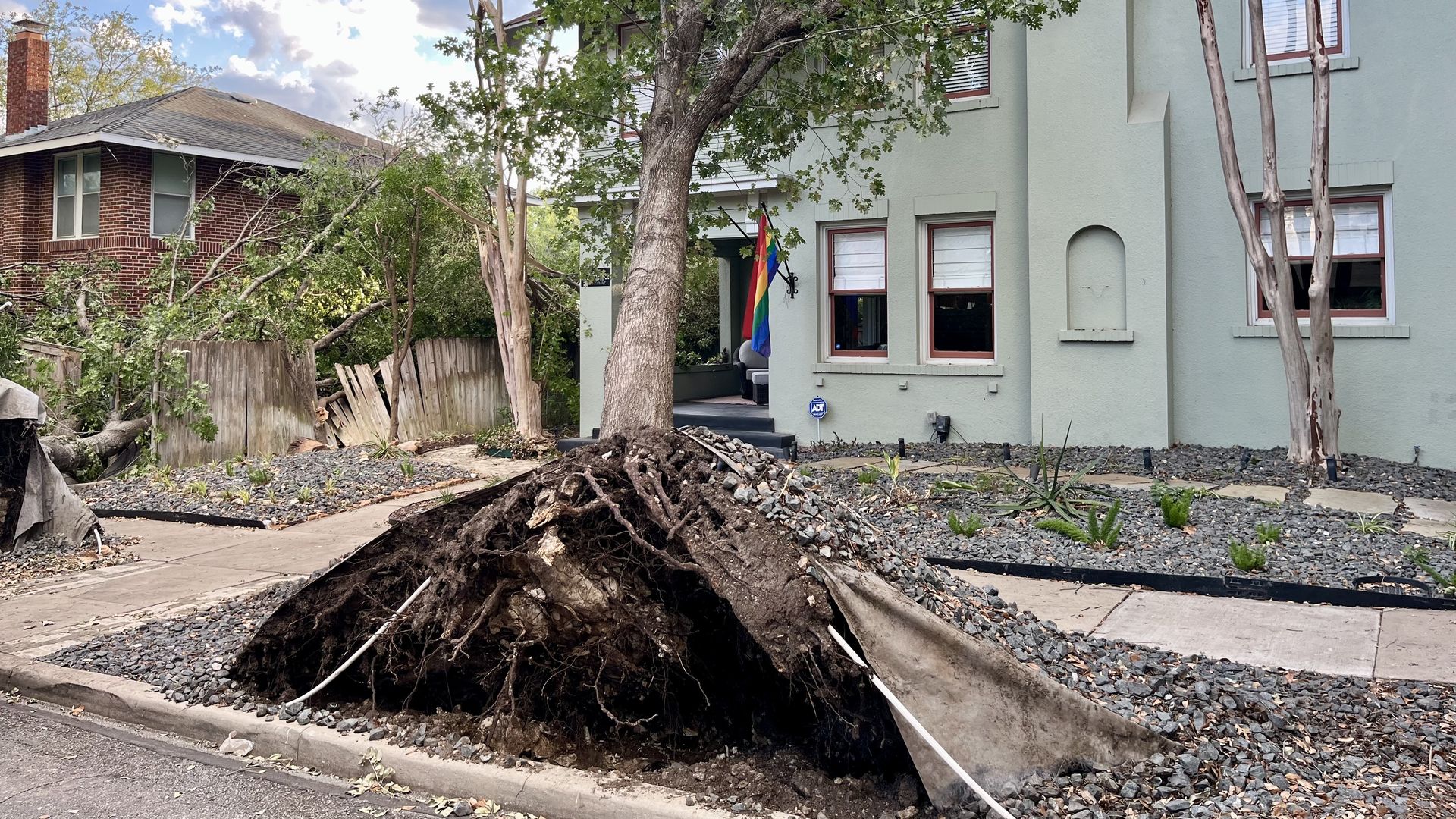 Uprooted tree with exposed roots and a large hole in the front yard, in front of a pale green two-story house with red-trimmed windows; a rainbow flag hangs nearby under a blue sky.