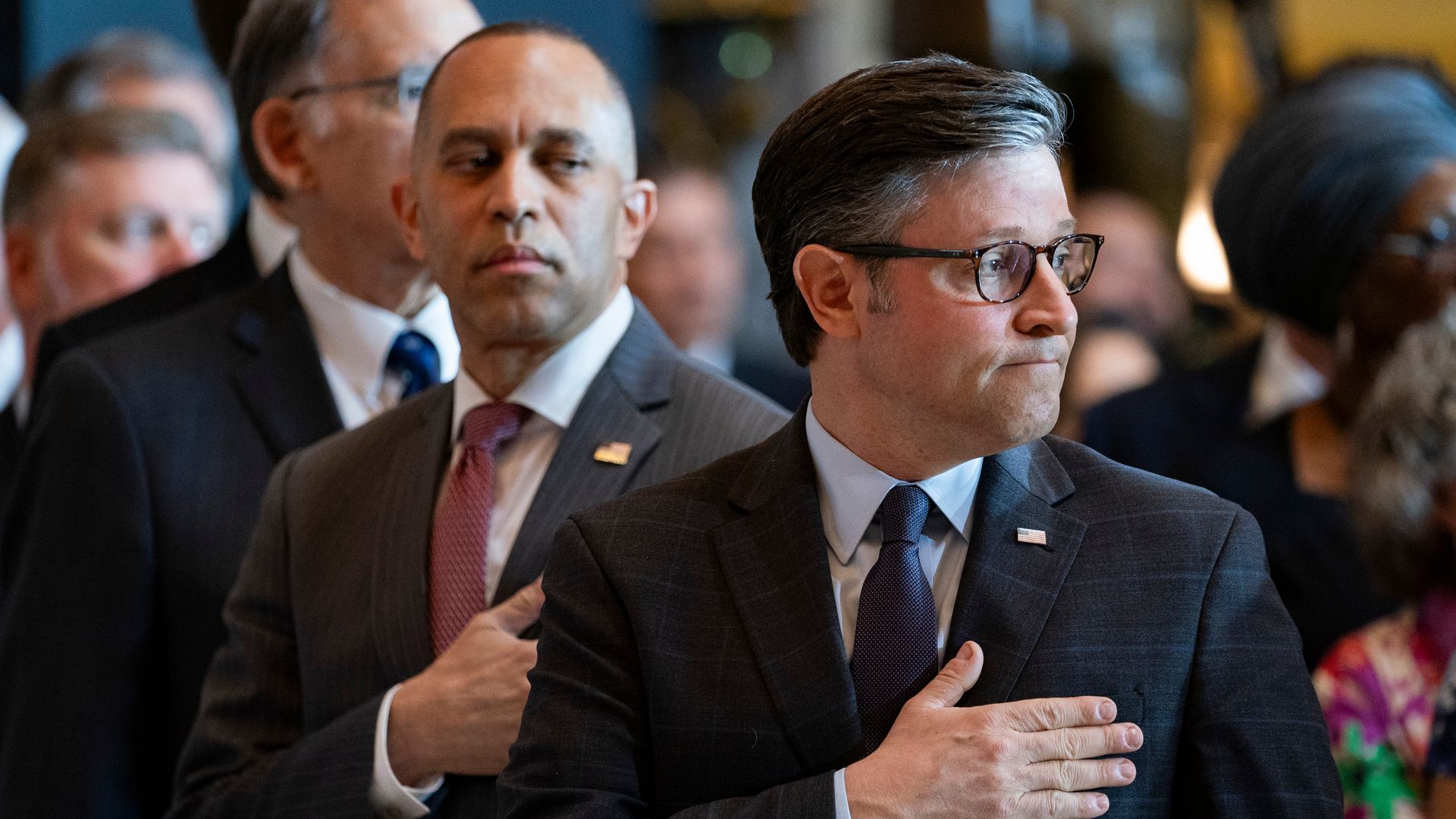 House Minority Leader Hakeem Jeffries, wearing a gray suit, white shirt and red tie, and Speaker Mike Johnson, wearing a blue suit, light blue shirt, blue tie and glasses, both holding their hands over their hearts while surrounded by other people.