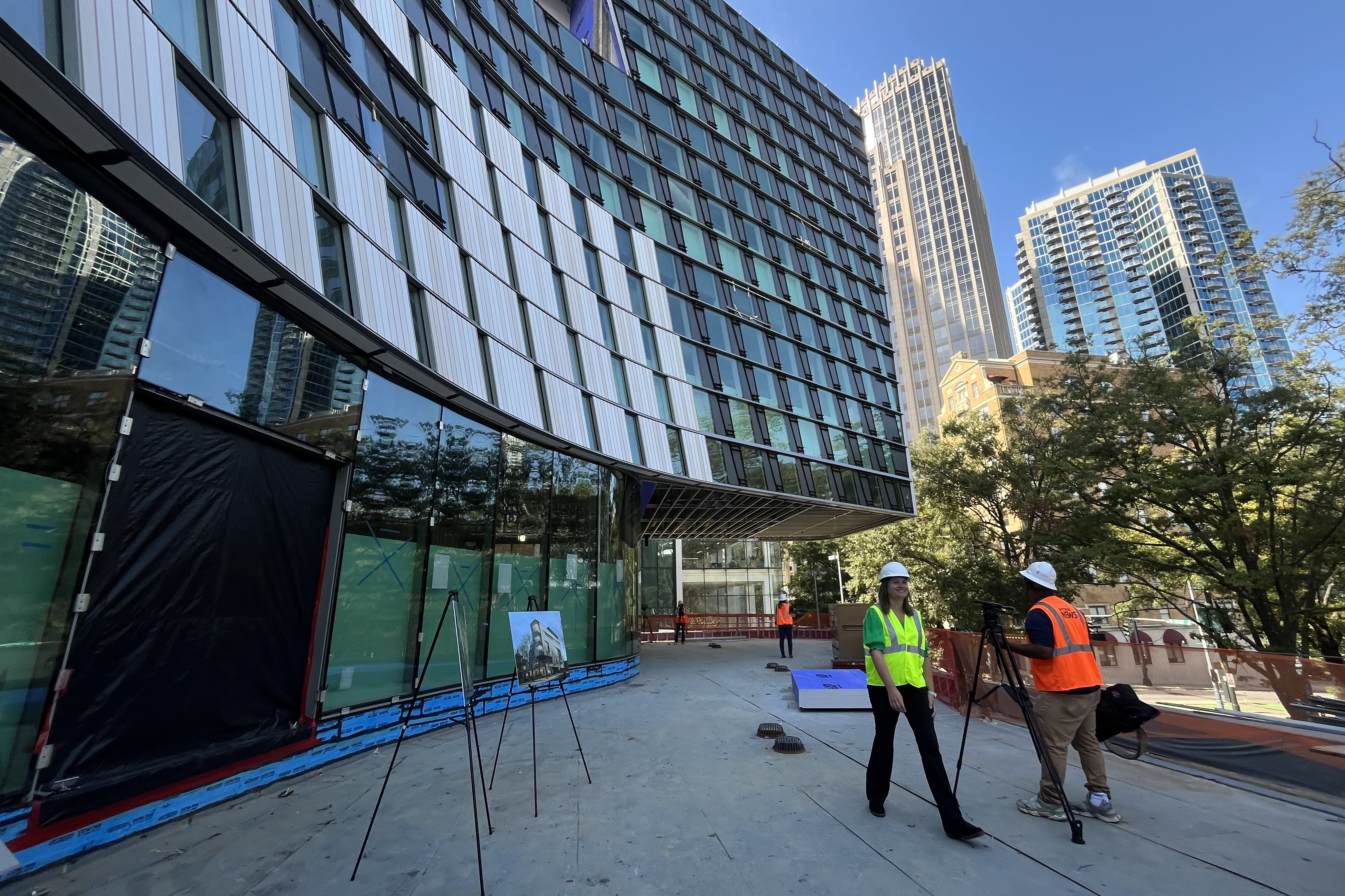 Curved modern building under construction with glass panels, two workers in neon vests and white helmets near a tripod camera, tall city skyscrapers and trees in the background.