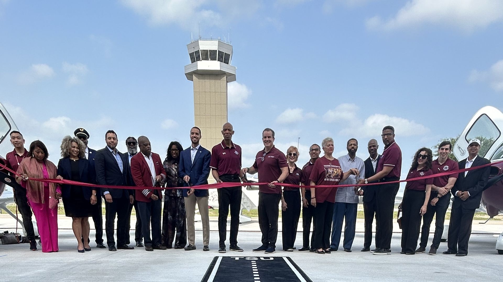 Group of people in maroon shirts and suits stand on an airport tarmac for a ribbon-cutting ceremony; a control tower looms in the background and a bold '27' runway mark is in the foreground.