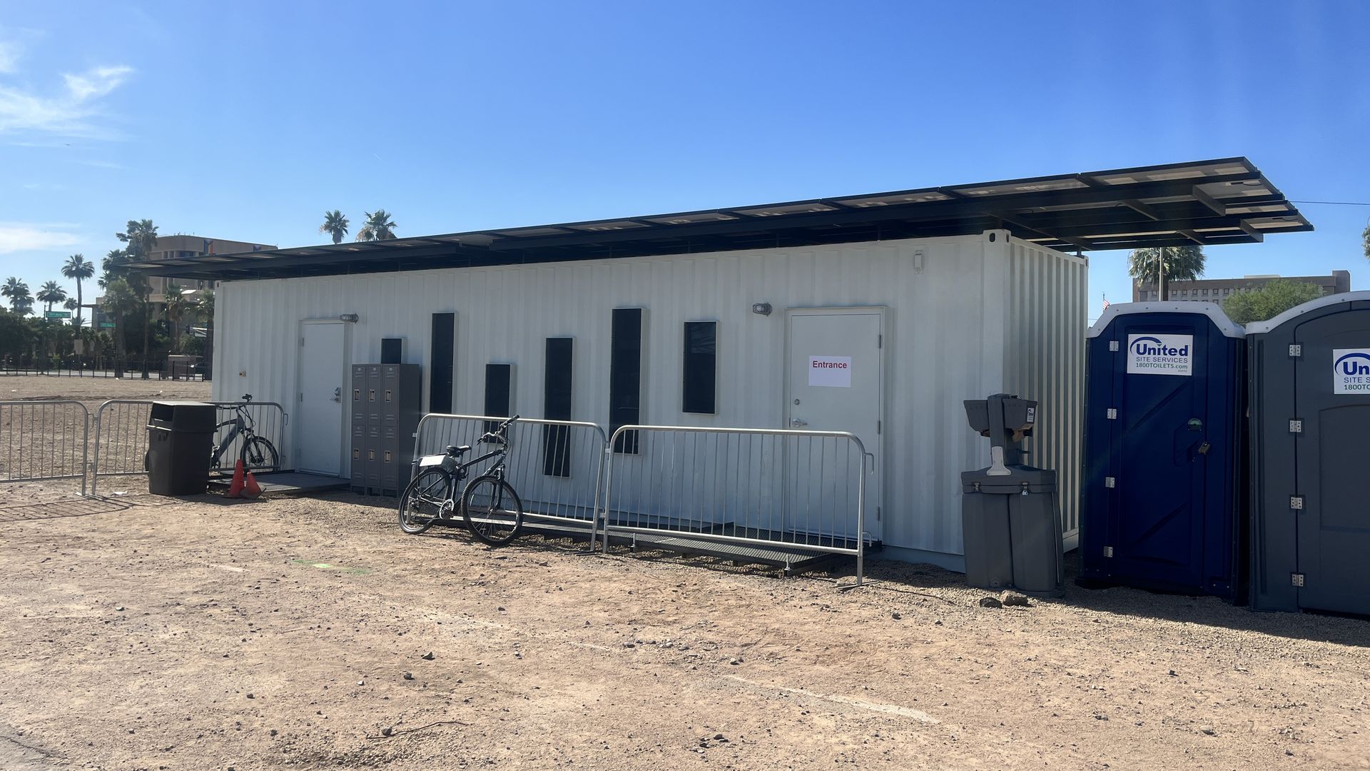 A shipping container with solar panels on the roof and adjacent port-a-potties.