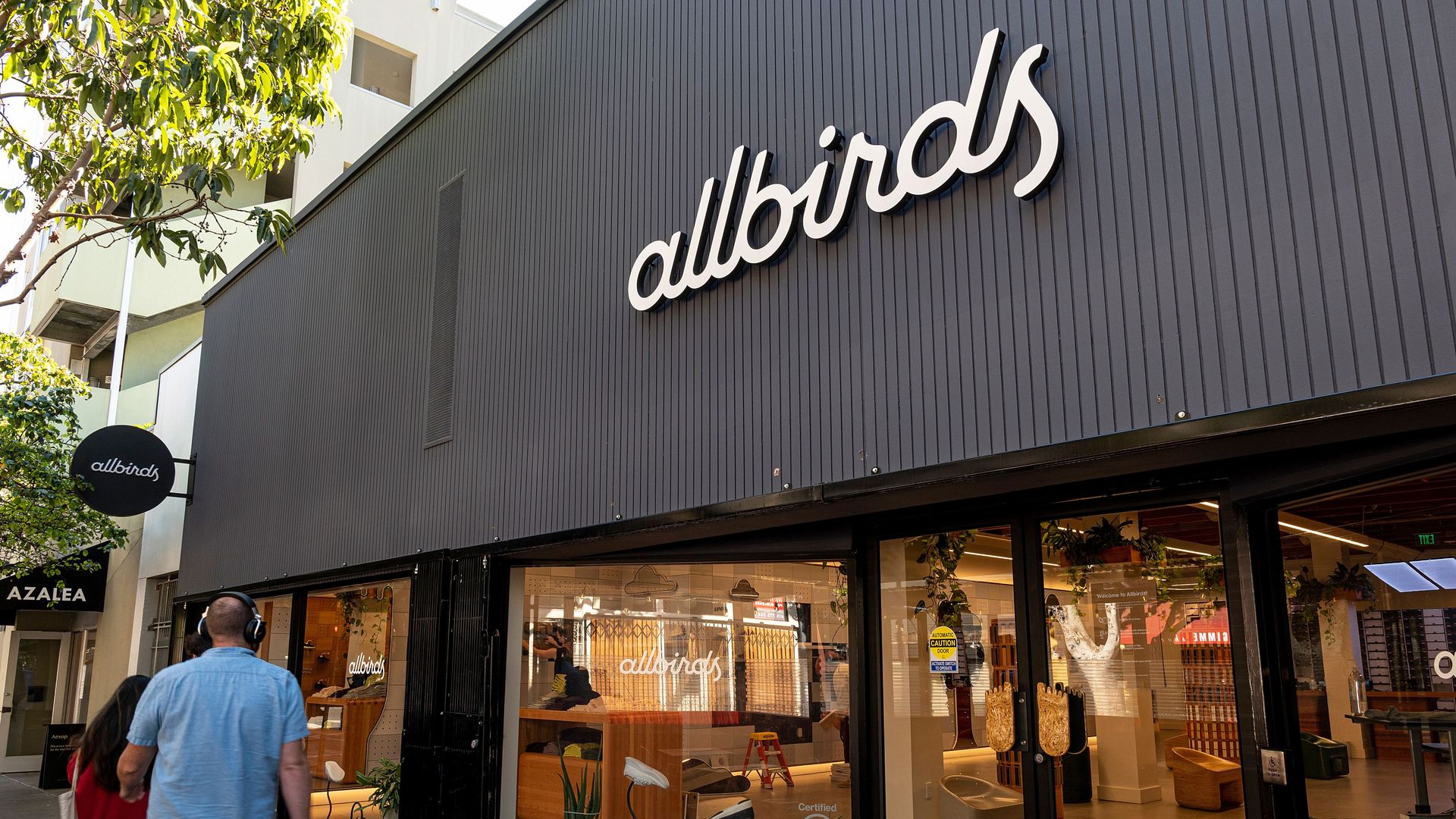 Storefront with a dark gray exterior and a large white "allbirds" sign. Glass windows show a bright interior with shoes and plants; a man in blue and a woman in red walk by.