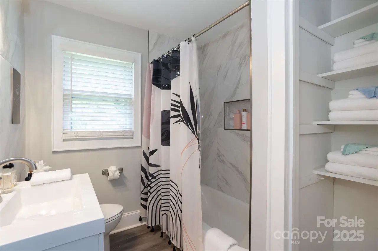 Bright bathroom with white sink, toilet, and window with blinds. Shower area has marble wall tiles and a black-and-white geometric patterned curtain. Open shelving with white towels.