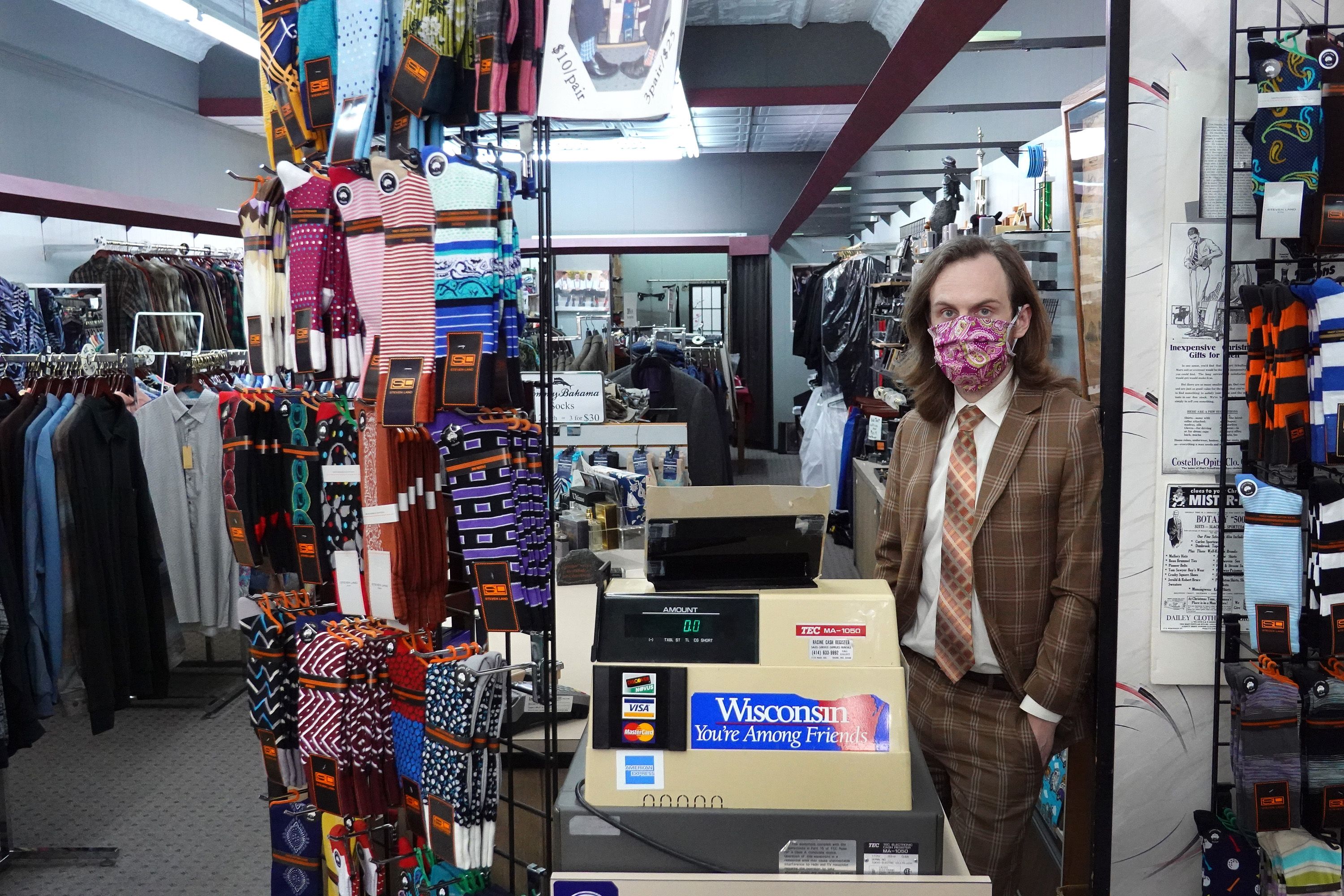 In this image, a man in a suit stands in a retail store surrounded by shirts