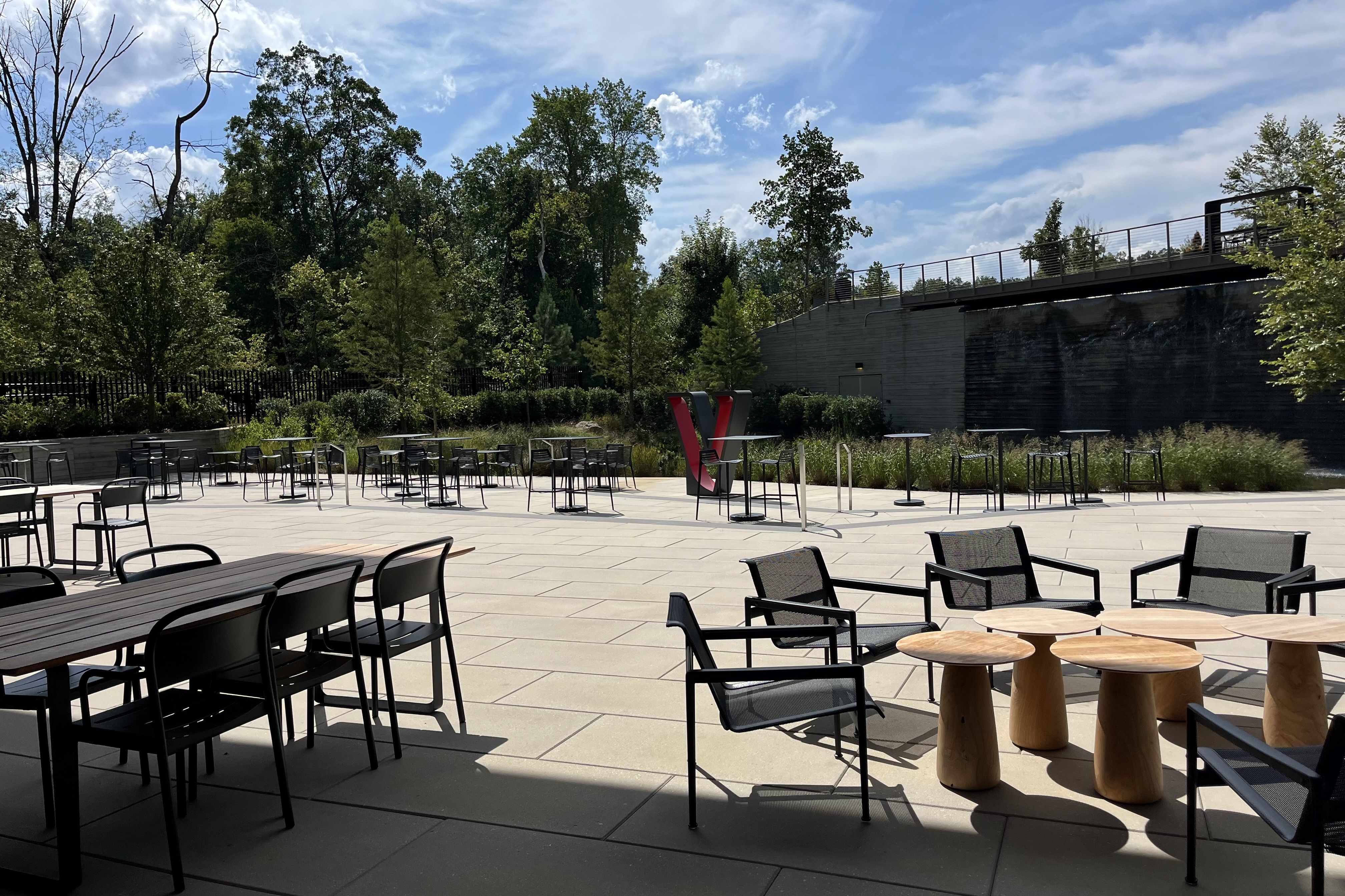 Outdoor patio with black chairs and wooden tables on light stone tiles under blue sky with scattered clouds and tall green trees surrounding the area.