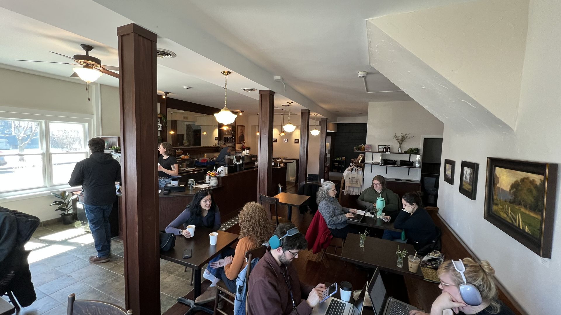 A coffee shop with brown wood beams, a bar, patrons sitting at tables and art on the walls