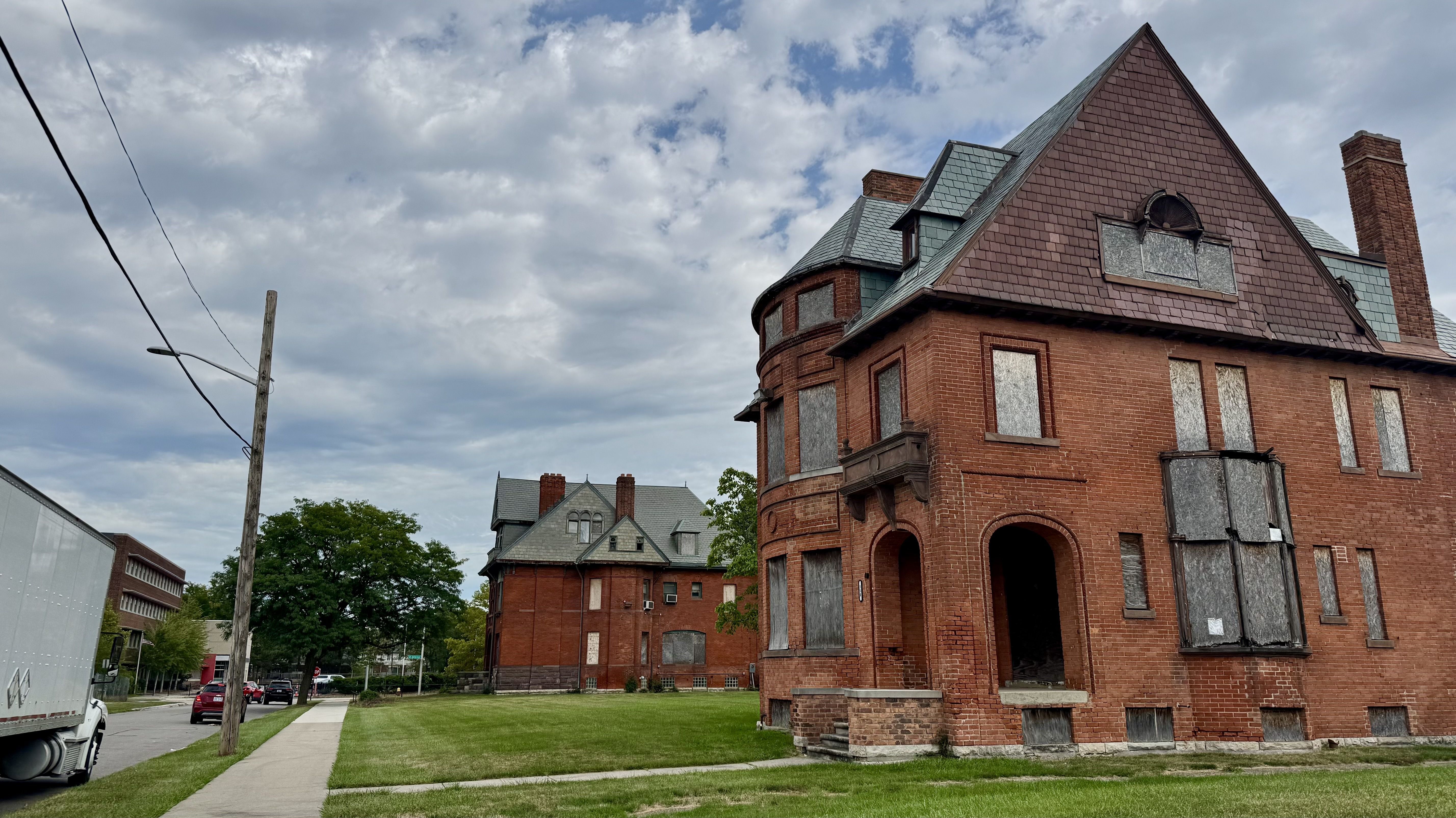 Large red brick building with a steep slate roof and rounded corner turret; boarded windows, arched entrance, and tall chimney. Lawn, sidewalk, power pole nearby beneath a cloudy sky.