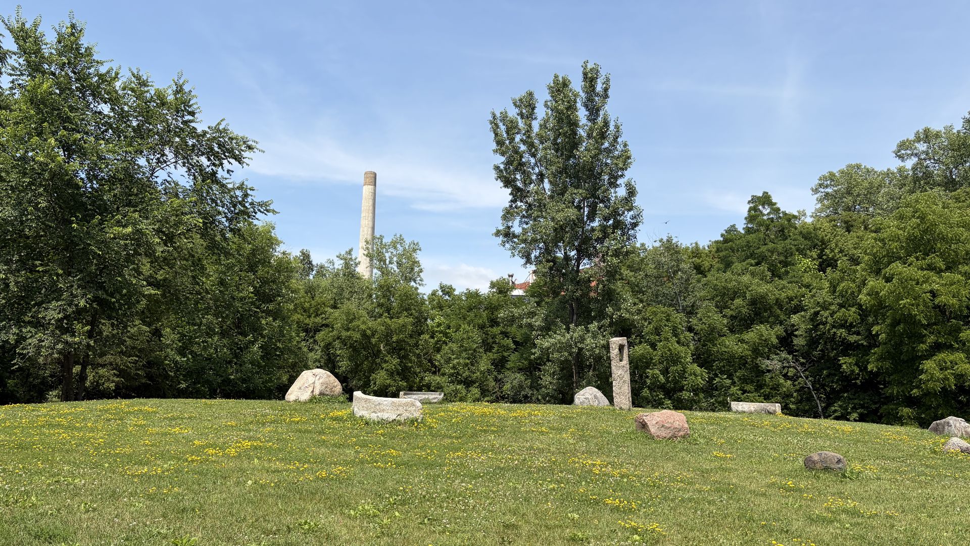 Bright park scene with a blue sky, dense green trees, a tall concrete chimney in the middle-left, and scattered rocks on a grassy field dotted with yellow flowers.