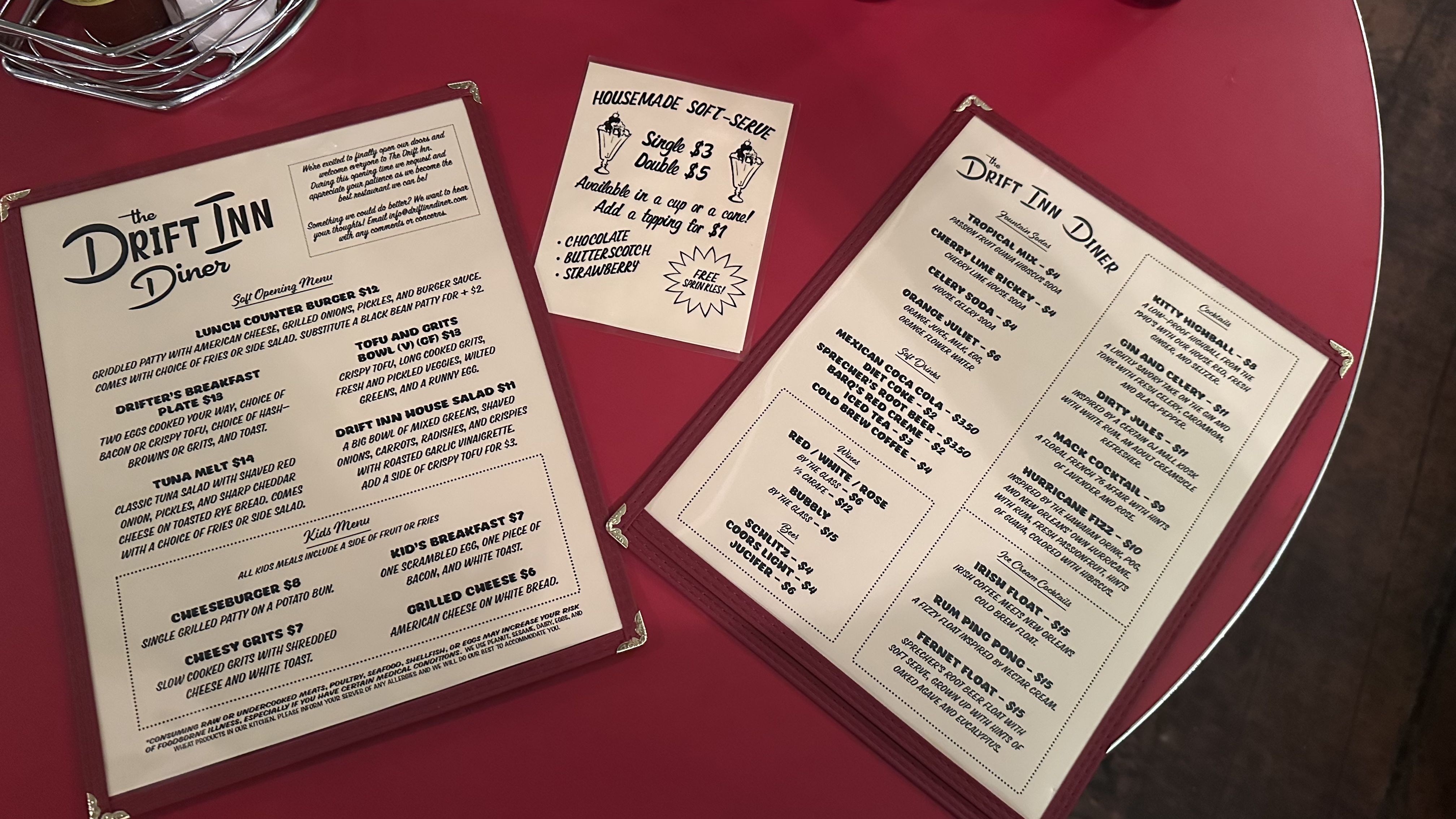 Two Drift Inn Diner menus on a red table: soft opening menu with burgers, breakfast, salads, kids options and a soft-serve menu with chocolate, butterscotch, strawberry flavors. Drink menu includes sodas, wines, beer, cocktails, and ice cream cocktails.