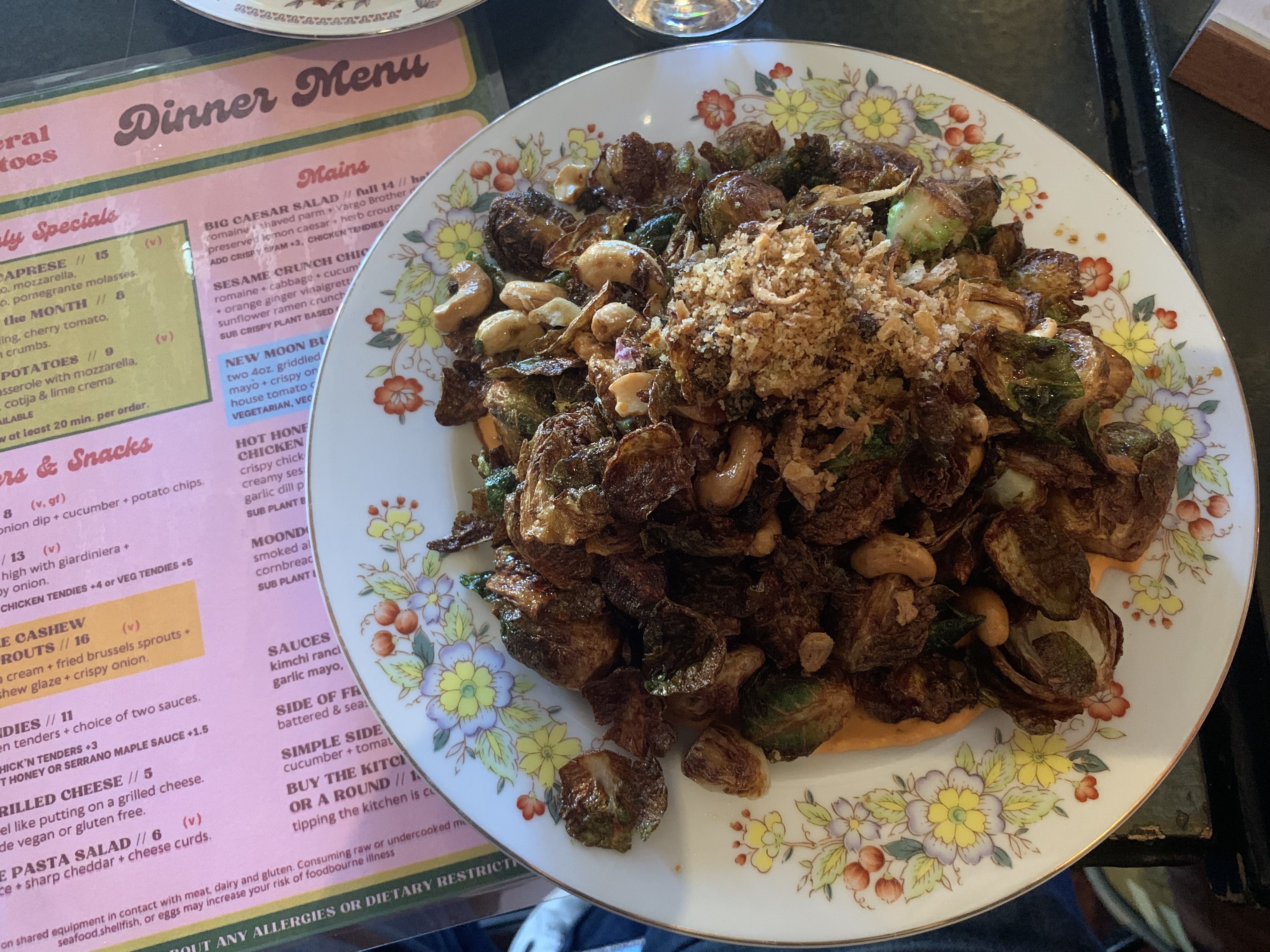 Plate of roasted vegetables, including brown Brussels sprouts and greens, topped with a crispy breadcrumb crust; cashews scattered nearby. Floral-patterned plate on a pink Dinner Menu sheet.