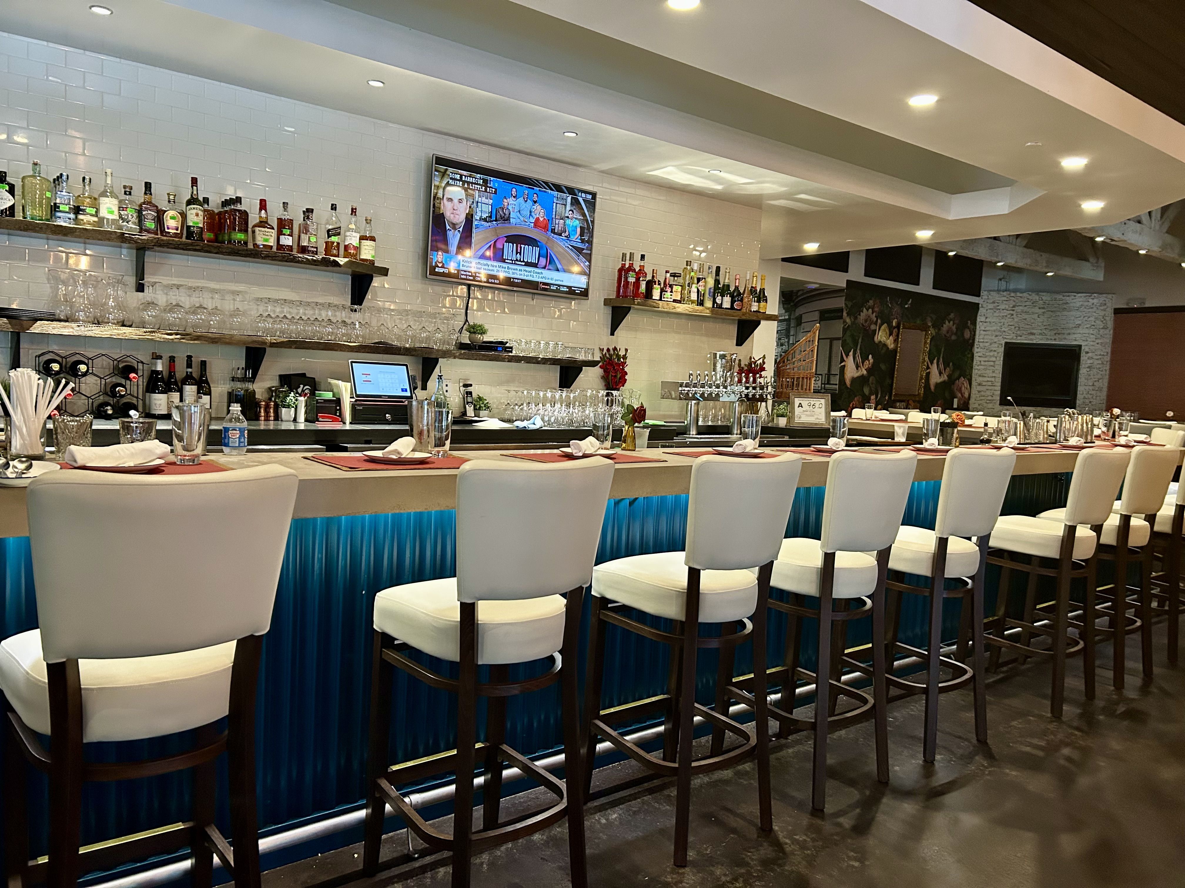 Modern bar with white cushioned stools, blue under-bar lighting, shelves of liquor bottles, a TV showing a sports program, and neatly set tables with glasses and napkins.