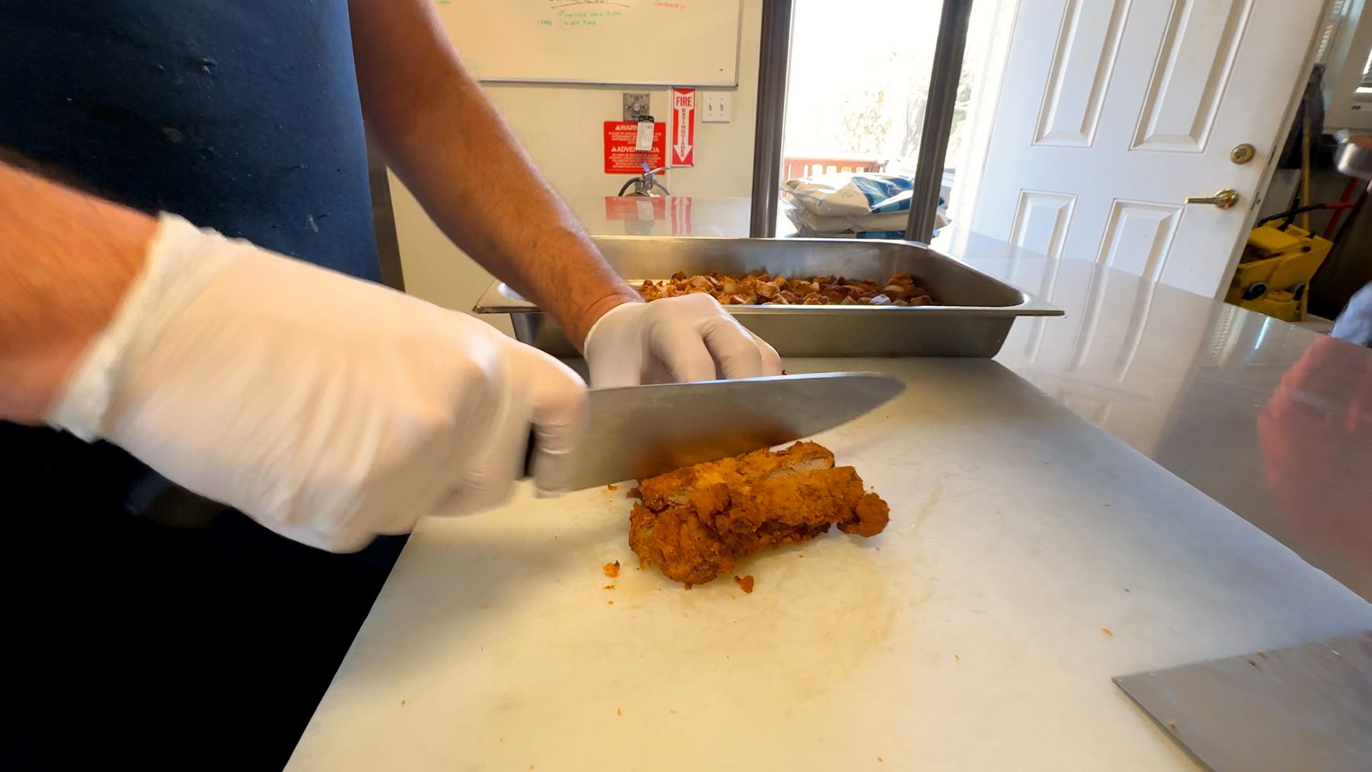 Person wearing white gloves slicing fried chicken on a white cutting board in a kitchen with a tray of diced chicken and a door in the background.
