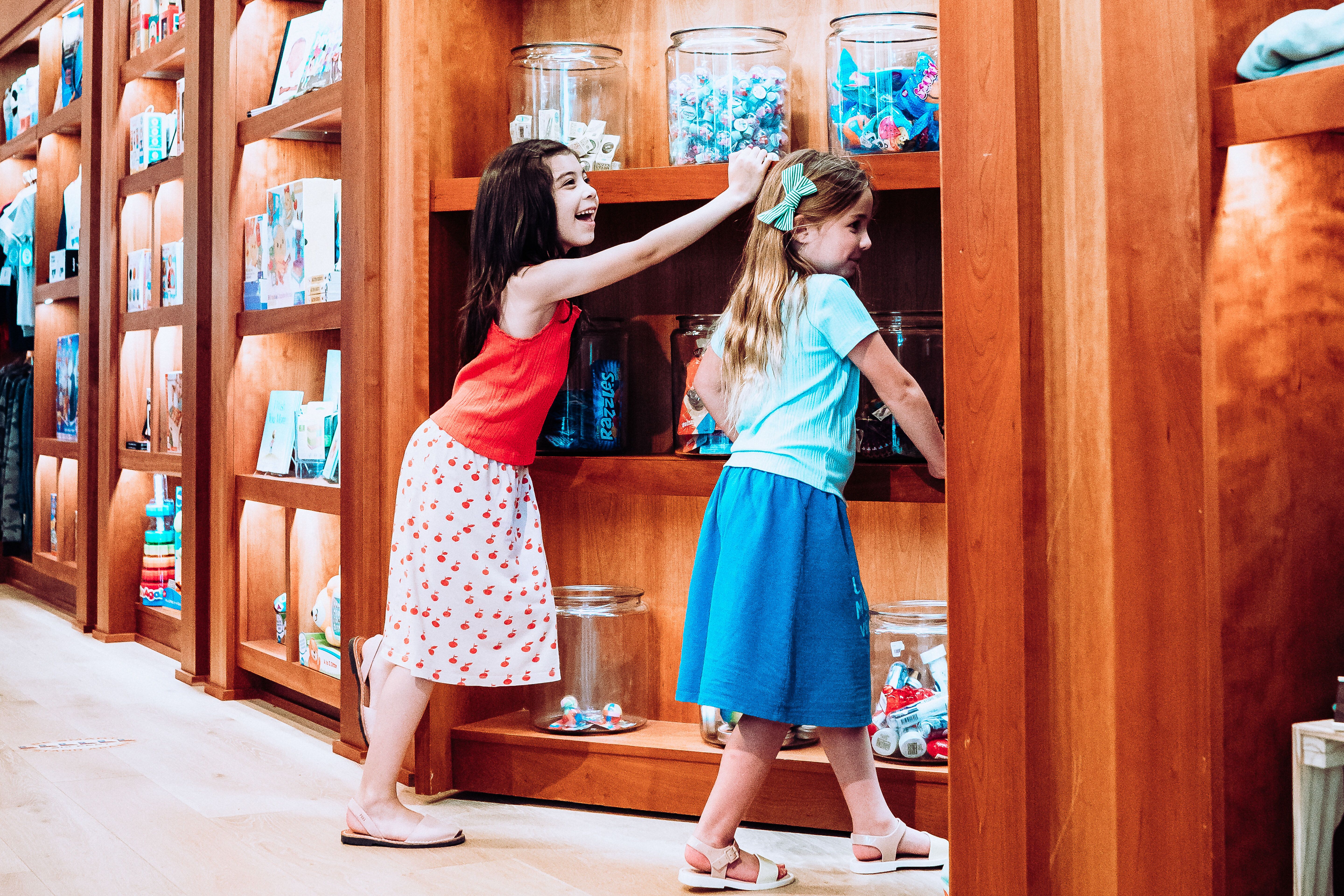 Two young girls in a store; one in a red top and white skirt with red patterns, the other in a light blue top and blue skirt, reaching into large glass jars on wooden shelves.