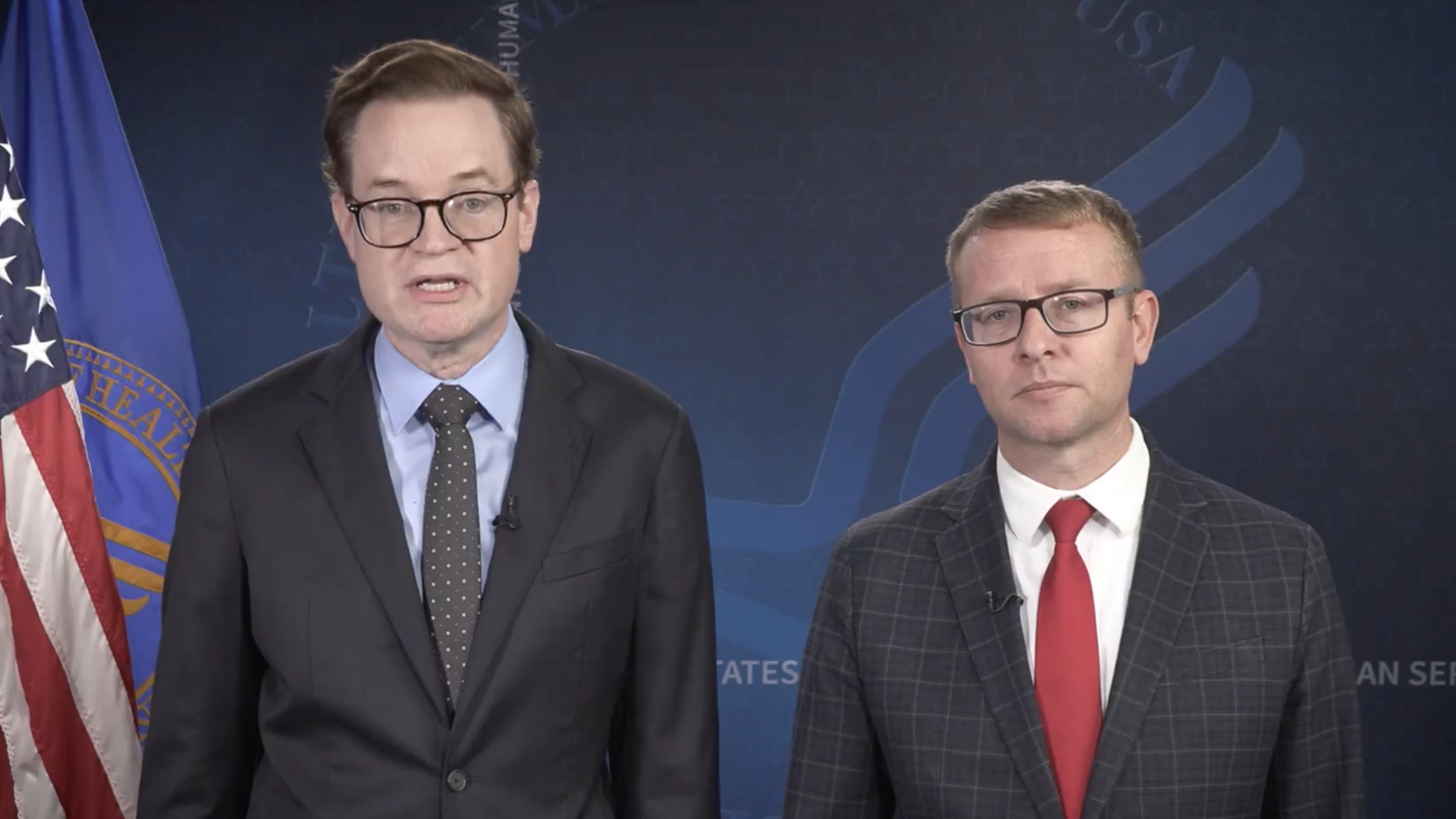 Two men wearing glasses and suits in front of U.S. and health department flags. HHS Deputy Secretary Jim O'Neill wears a dark suit with a blue shirt and polka dot tie and ACF Assistant Secretary Alex Adams wears a plaid suit with a red tie.
