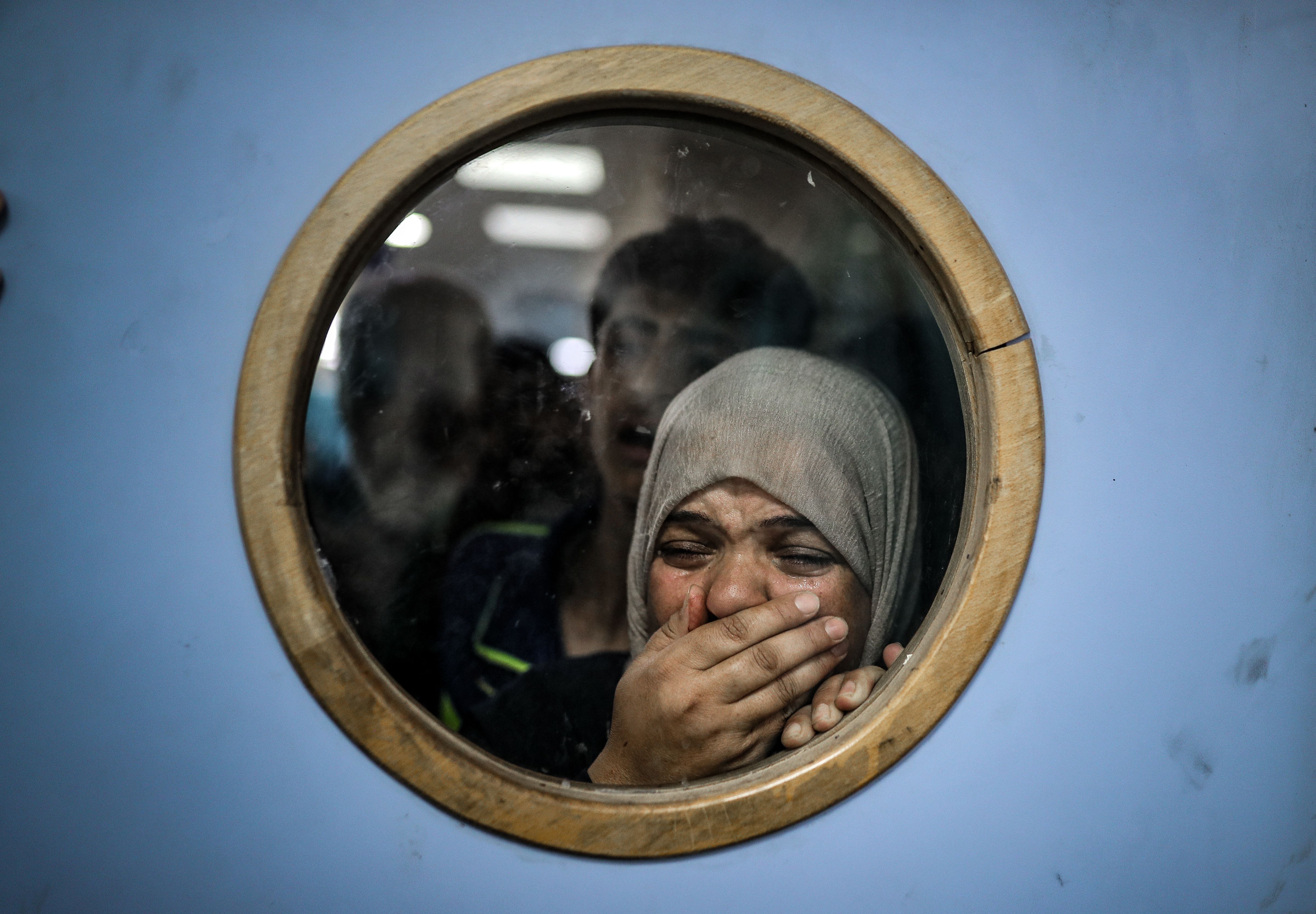 A woman looks through a hospital door window, crying and covering her mouth. 