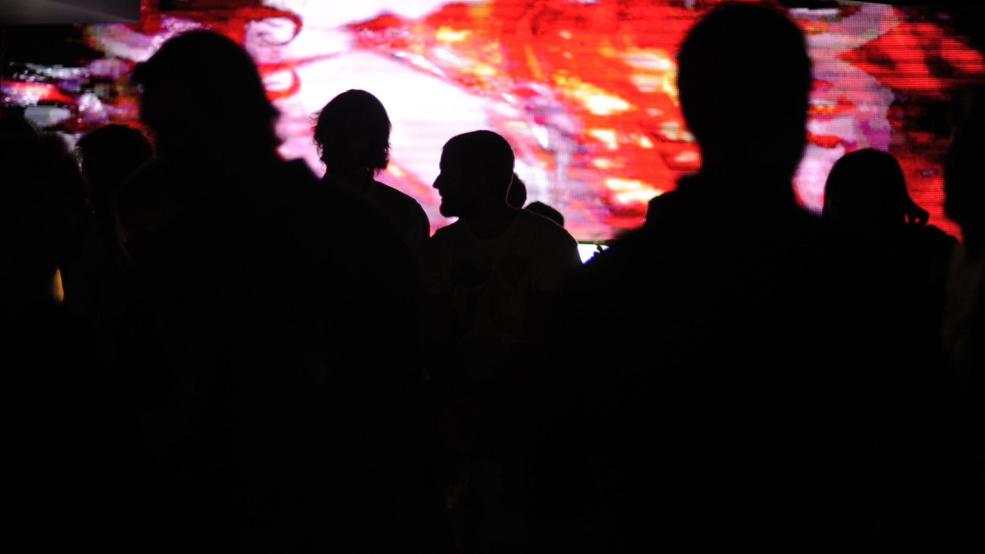 Silhouetted group standing in a dark room, facing a large bright red-pink abstract light display that fills the background with vivid colors.