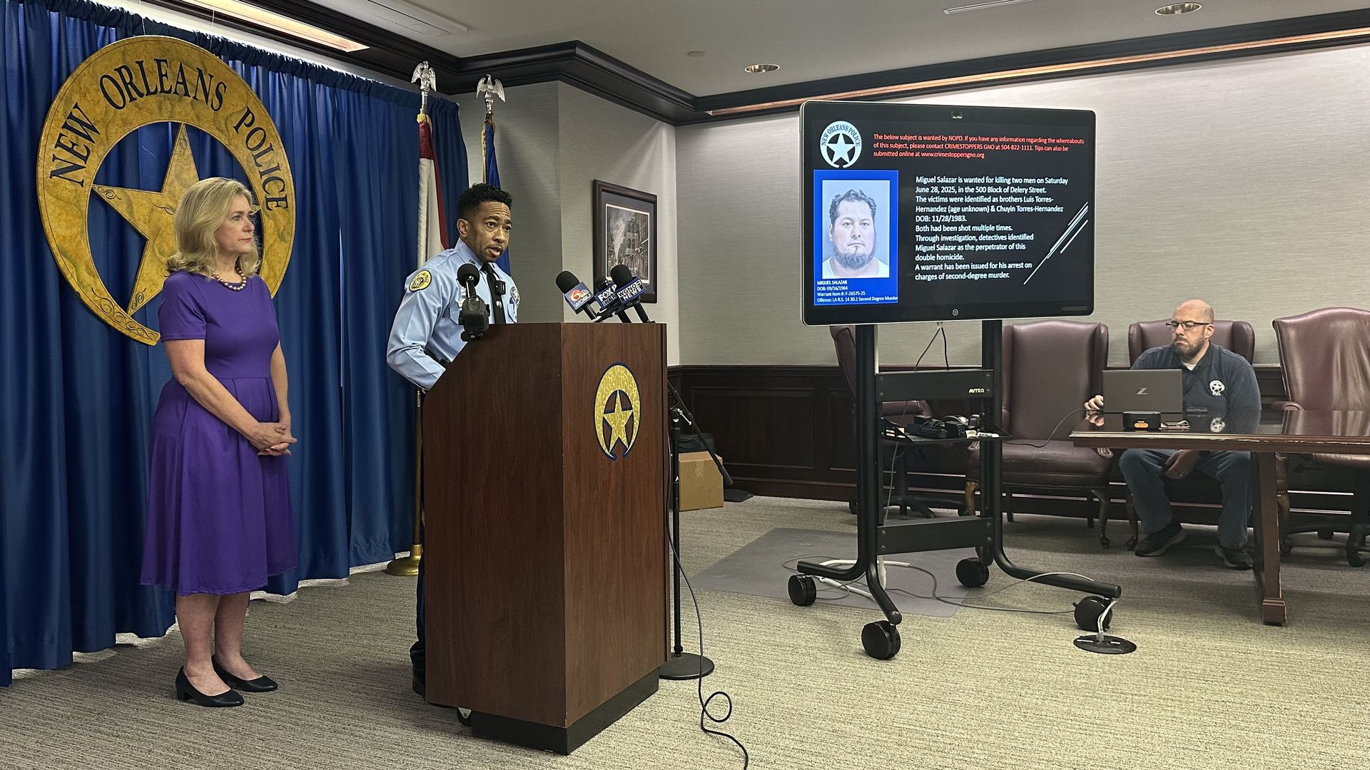 A police officer speaks at a New Orleans Police press conference with a woman in a purple dress standing nearby and a man sitting at a table with a laptop, a suspect's photo on a screen.