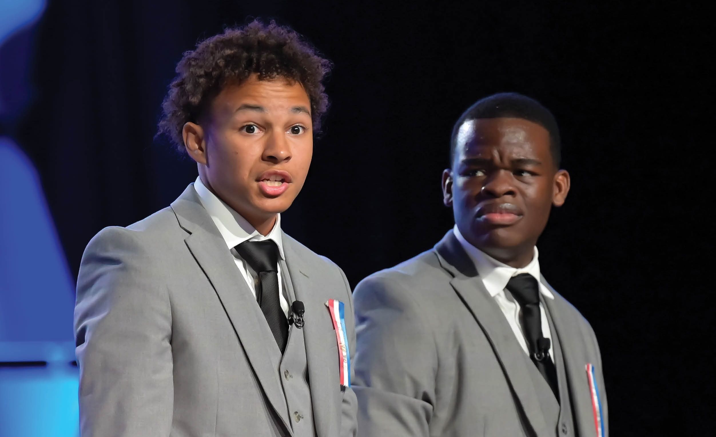 Two young men in grey suits and black ties with lapel microphones perform a dramatic piece together