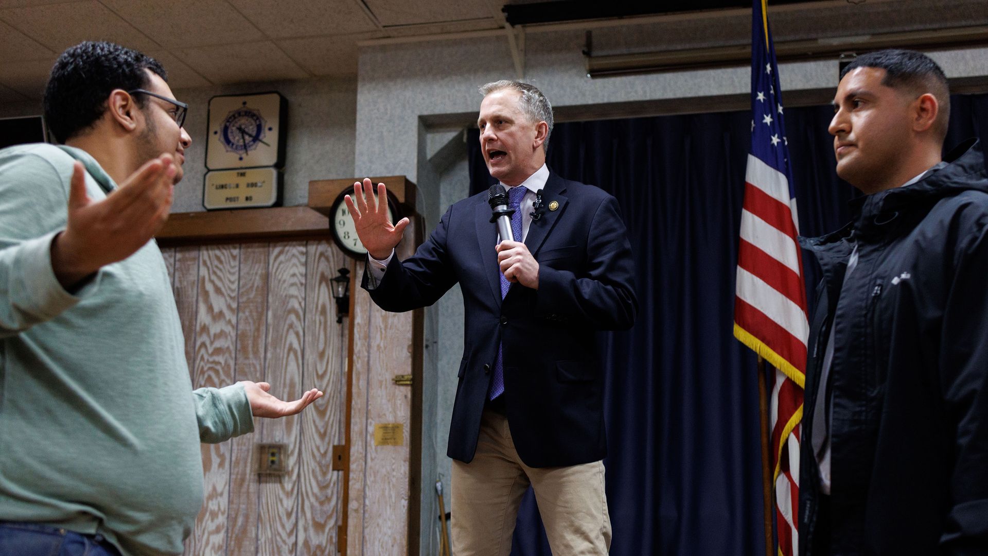 Rep. Sean Casten, wearing a blue suit and khakis, holds up his hand at a man in a light green shirt while standing on a stage in front of a blue curtain, a wooden wall and an American flag.