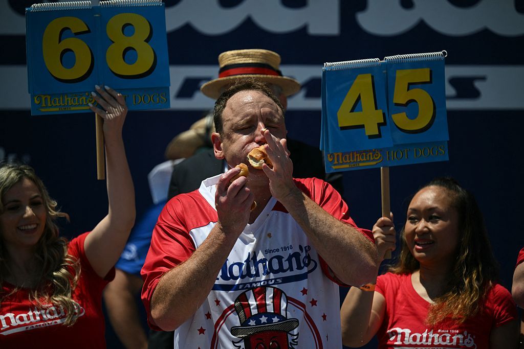 A photo showing Joey Chestnut shoving a hot dog into his mouth. Behind him, two women hold signs with numbers: 68 and 45.