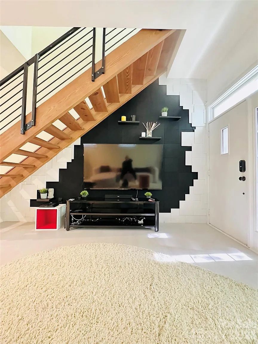 Modern living room with wooden staircase, black and white wall design behind a flat-screen TV, black TV stand, small plants, and a round beige rug near the white front door with a vertical window.