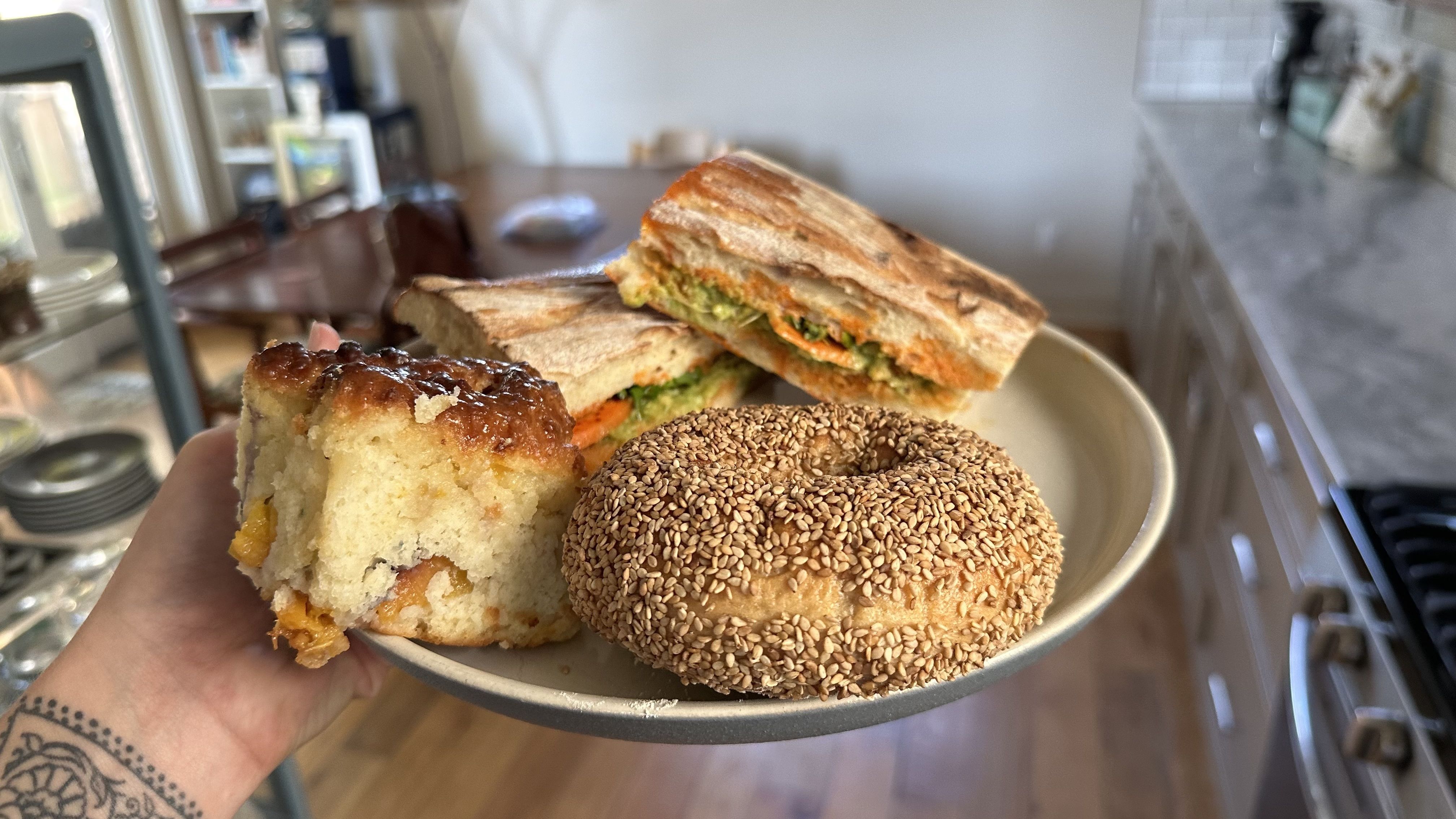 A hand holds up a plate filled with a bagel, a sandwich and a peach biscuit.