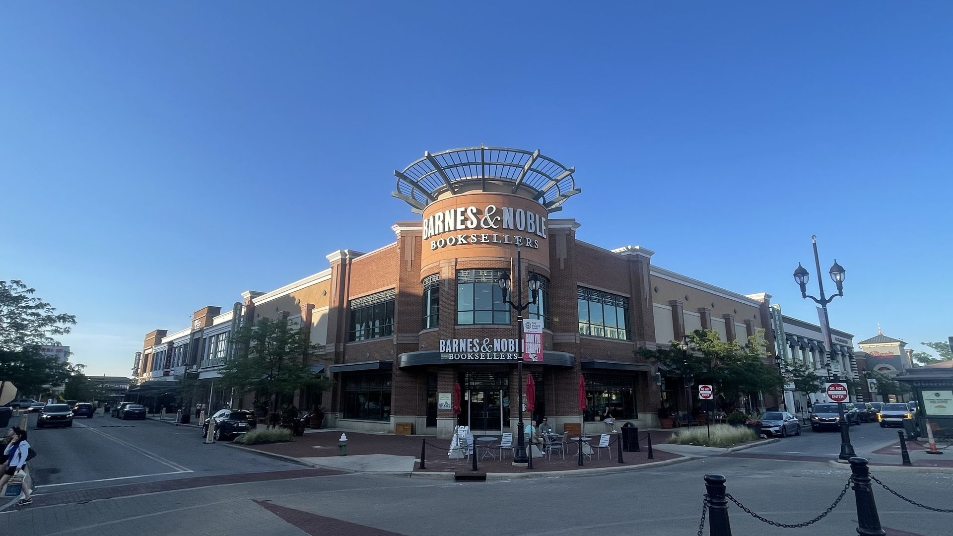 Exterior of Barnes & Noble Booksellers in a brick shopping plaza. A circular sign reads BARNES & NOBLE BOOKSELLERS beneath a metal arched canopy; blue sky, cars and pedestrians on the street.