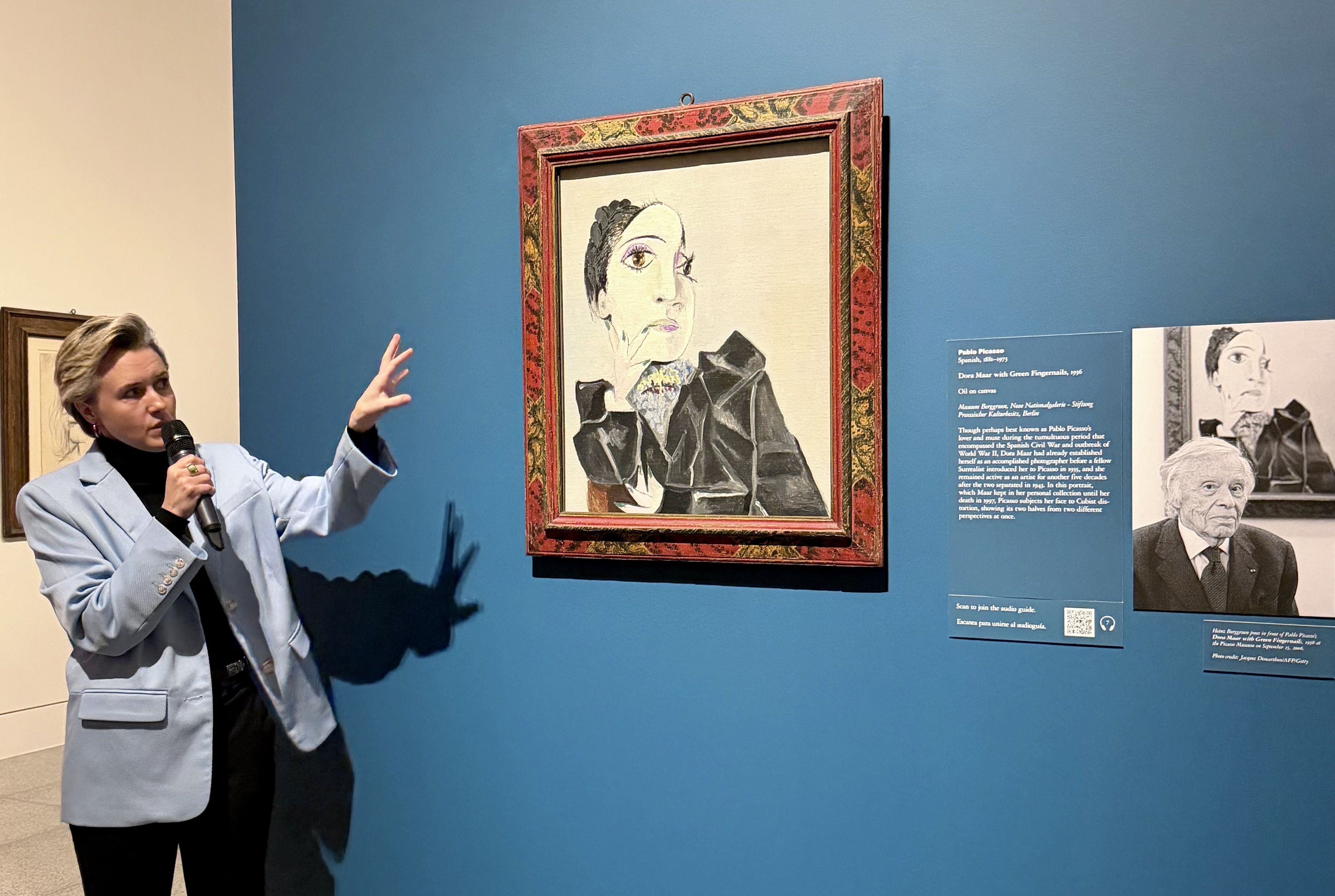 A woman in a light blue blazer speaks into a microphone, gesturing toward a framed Picasso portrait mounted on a blue gallery wall, with informational panels nearby.