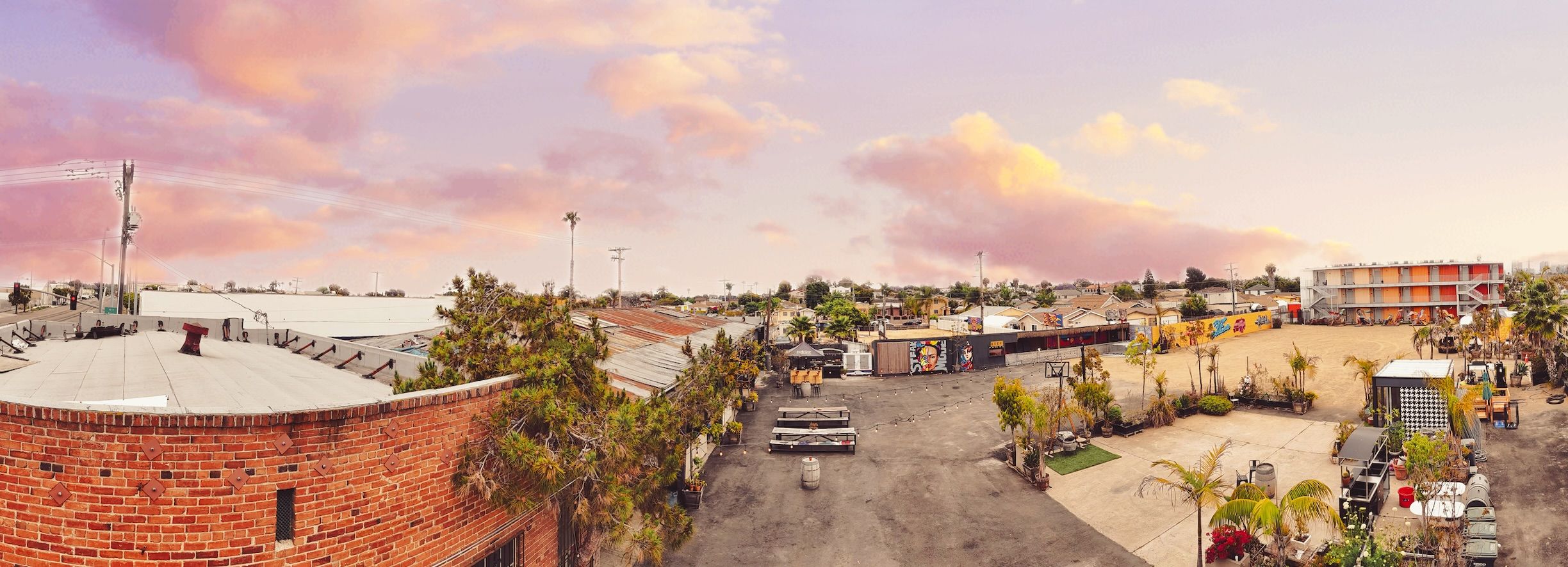 Panoramic view of an urban courtyard with brick buildings, picnic tables, palm trees, colorful graffiti walls, and a pastel purple and pink sky at sunset.