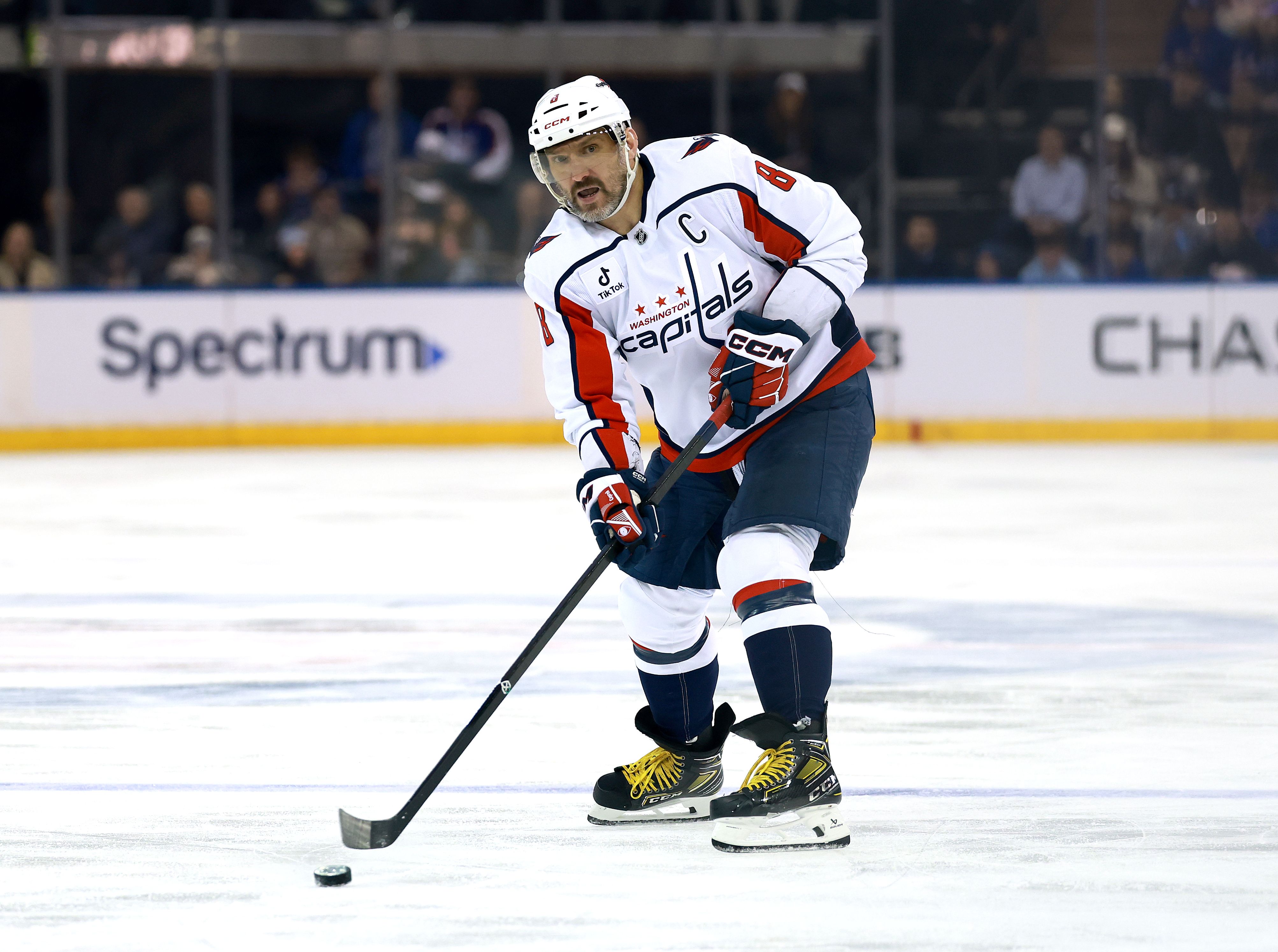 Washington Capitals captain Alex Ovechkin skates on the ice in a white jersey with red and navy trim, handling a hockey stick as the puck sits nearby; arena boards read Spectrum in the background.