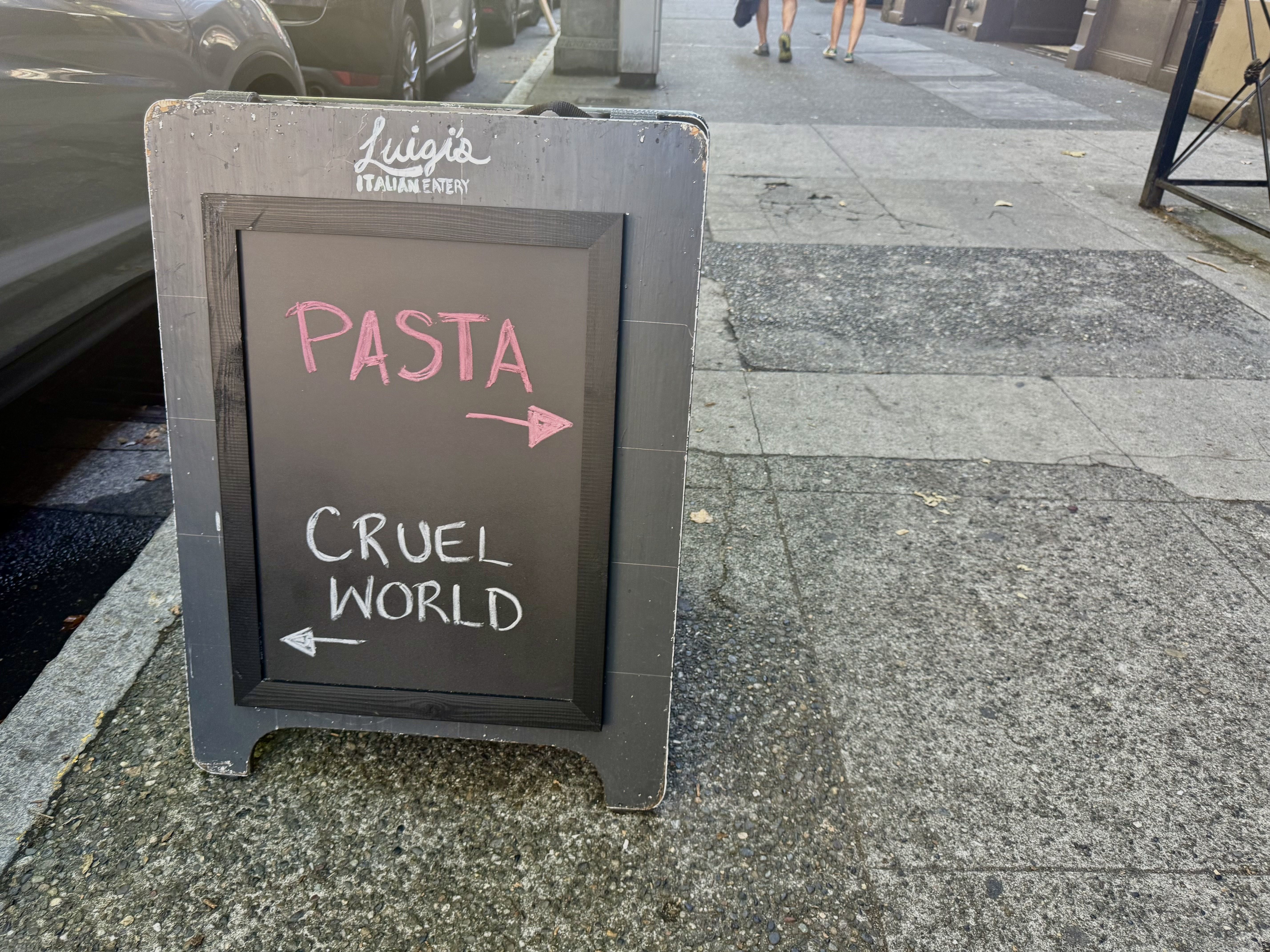 Sidewalk chalkboard sign for Luigi's Italian Eatery with pink arrow pointing right to "pasta" and white arrow pointing left to "Cruel World" on a gray sidewalk.