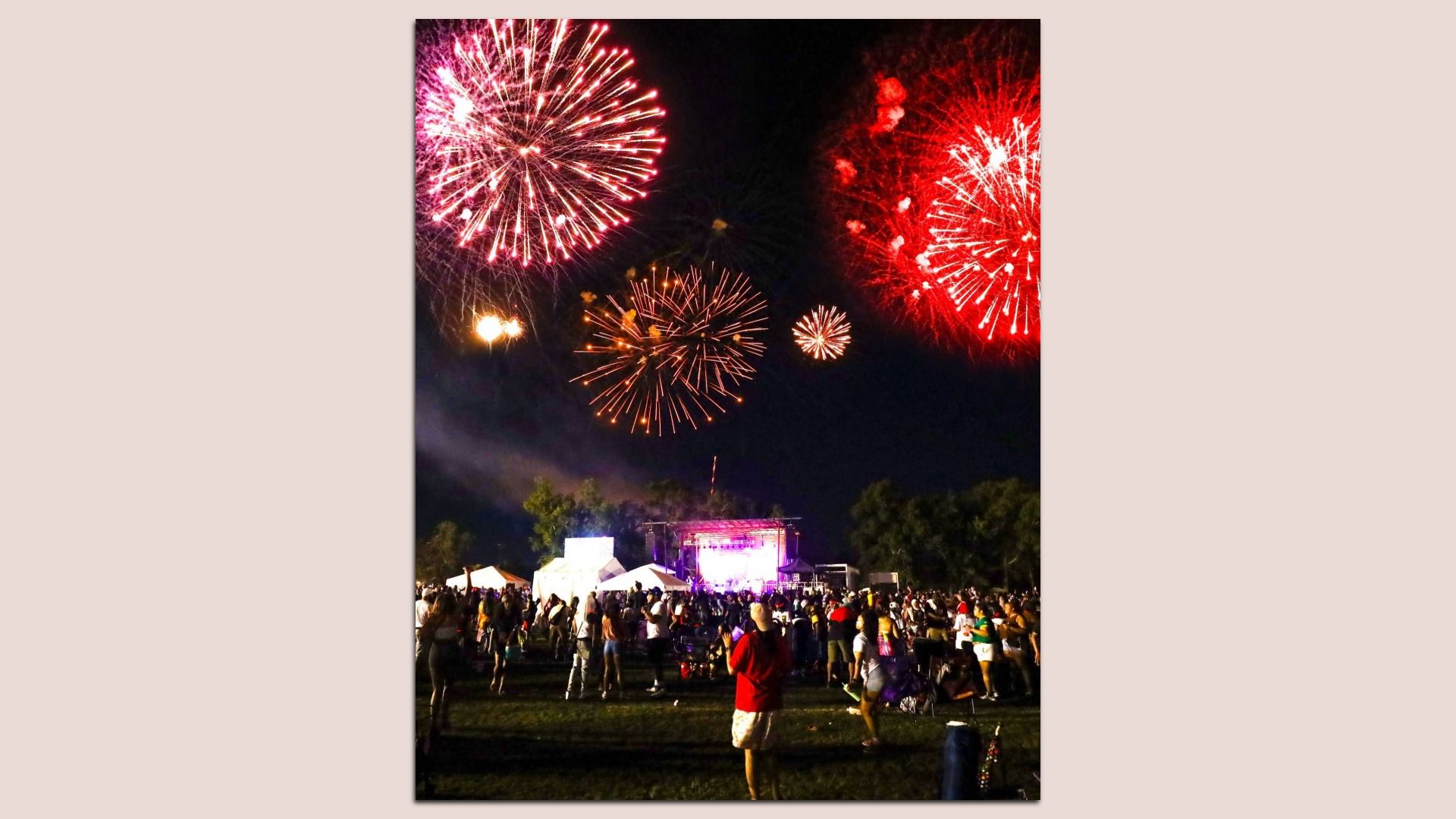 Night scene of a crowded outdoor festival with a lit stage and tents at the bottom; red and pink fireworks burst across the dark sky above.