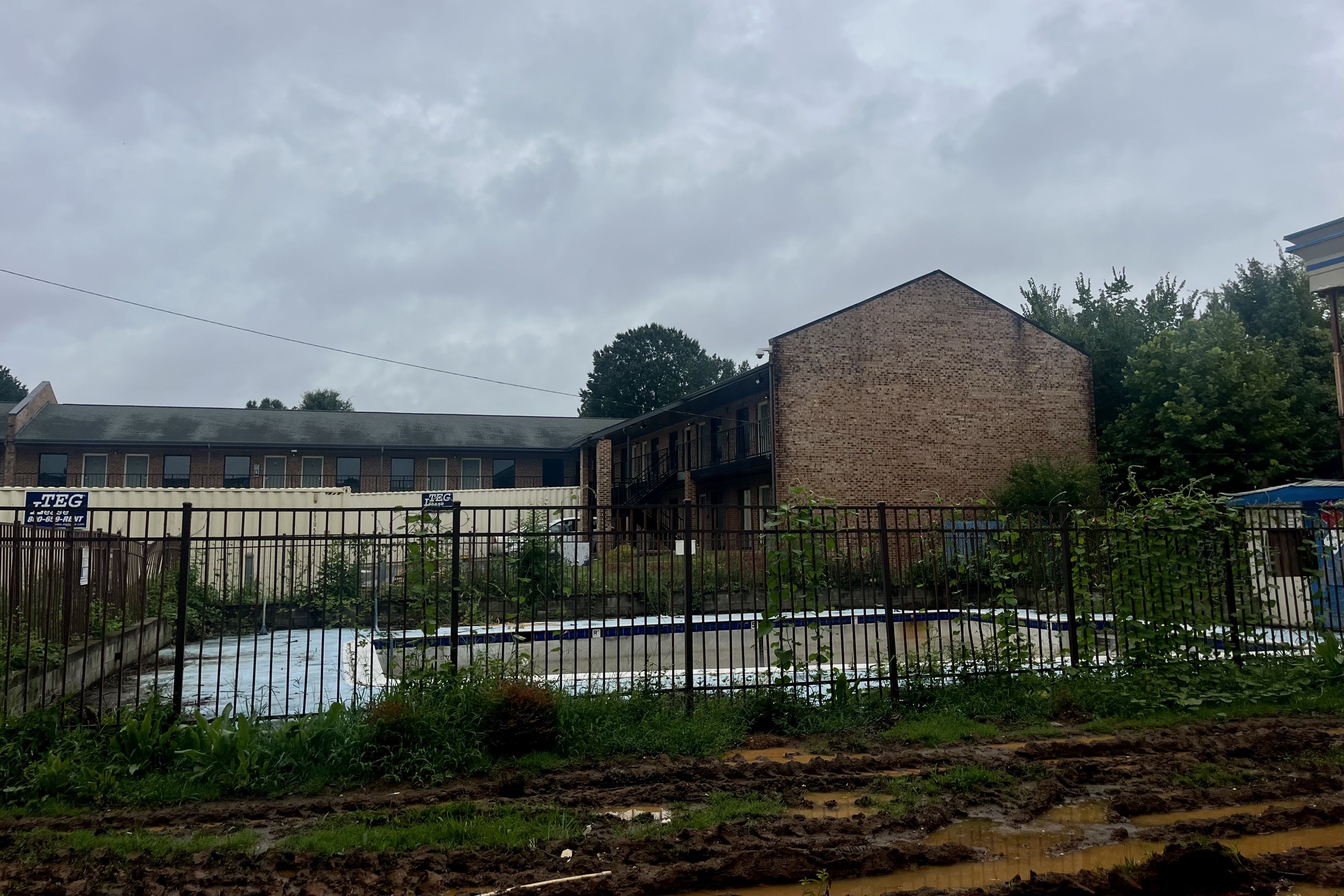 Empty, fenced outdoor swimming pool with overgrown grass and muddy ground in front, adjacent to a two-story brick building under a cloudy sky.