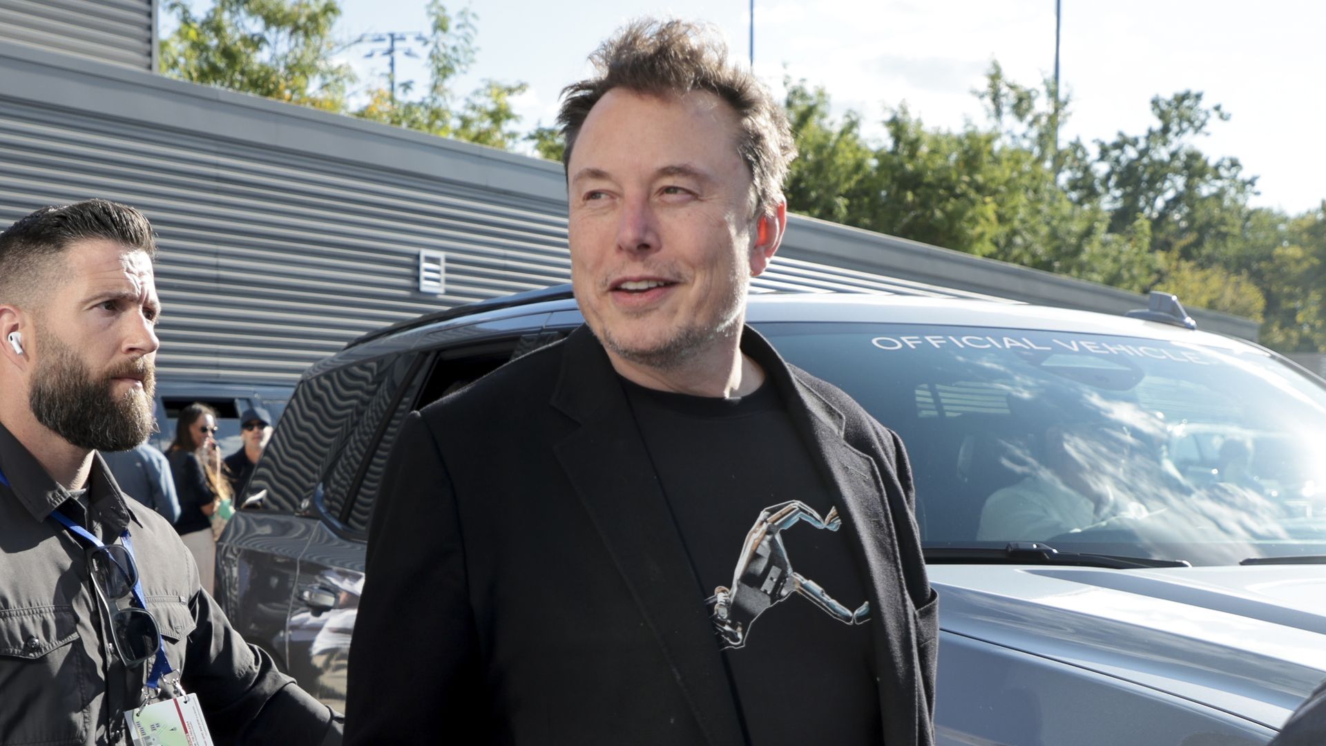 Elon Musk, dressed in a black t-shirt and jacket, stands by a car, as the wind blows back his hair, as he attends the 2024 US Open Tennis Championships at the USTA Billie Jean King Tennis Center on September 8, 2024 in Flushing Meadows, Queens, New York City.