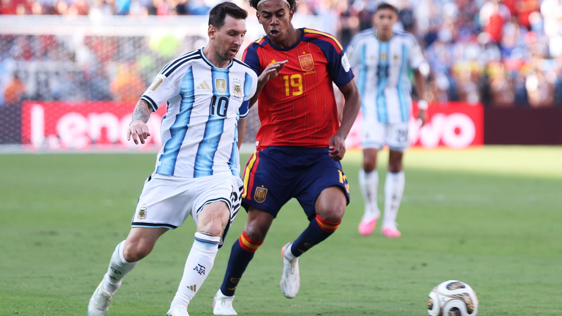 A soccer player in Argentina's white-and-sky-blue striped jersey dribbles the ball while a Spain player in a red jersey closely defends him during a match. Another Argentina player and a blurred crowd fill the background.